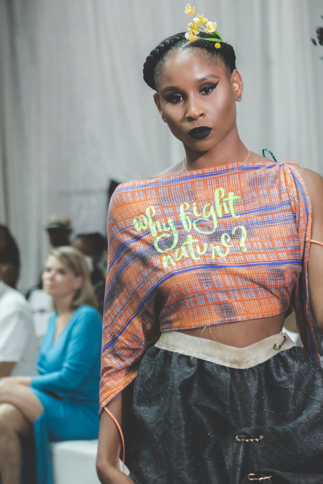 Young woman with braided hair and dark lipstick wearing a colorful top with text, standing indoors.