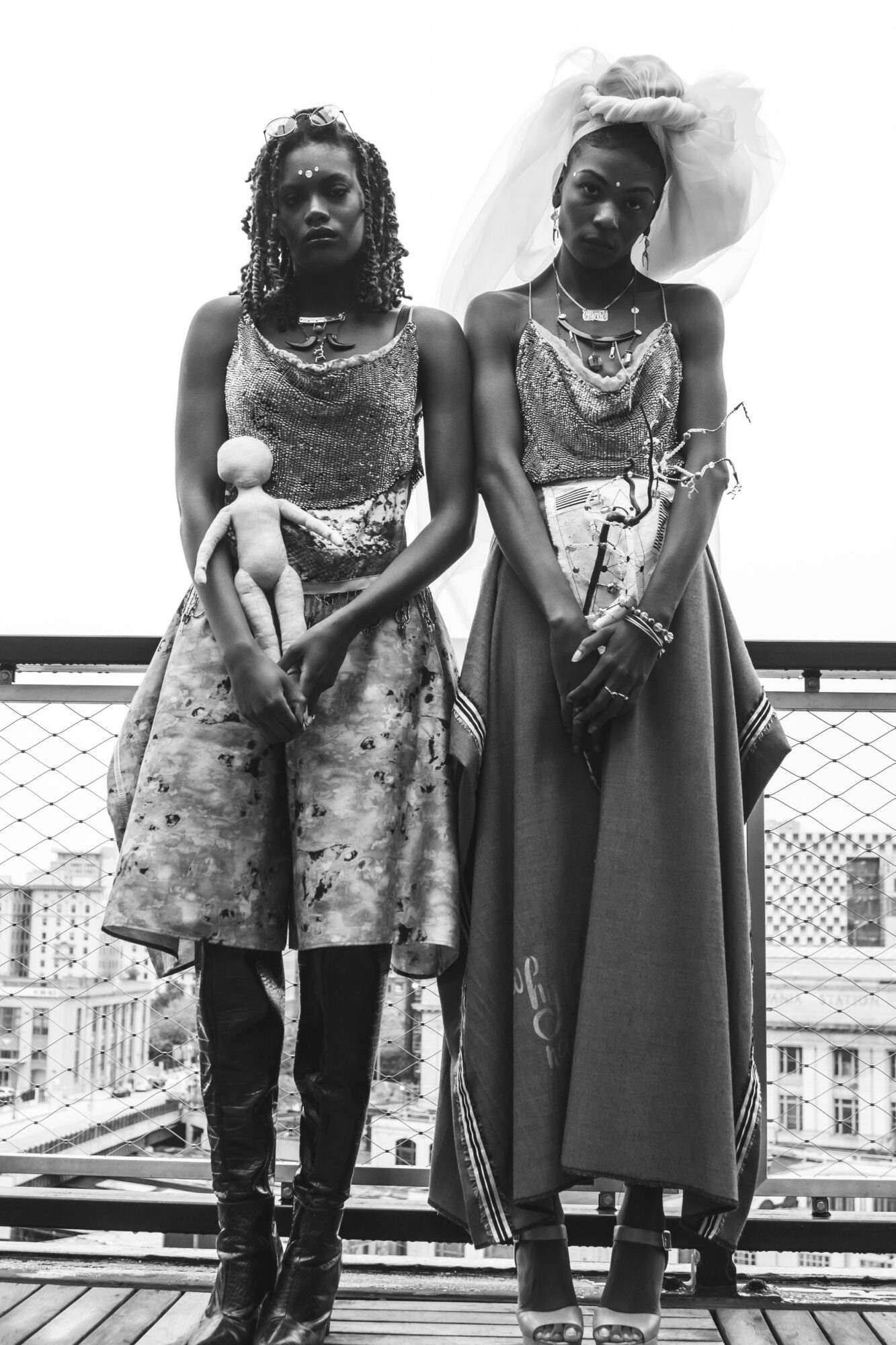 Two women stand on a balcony with city buildings in the background, wearing traditional clothing and jewelry, holding a doll.