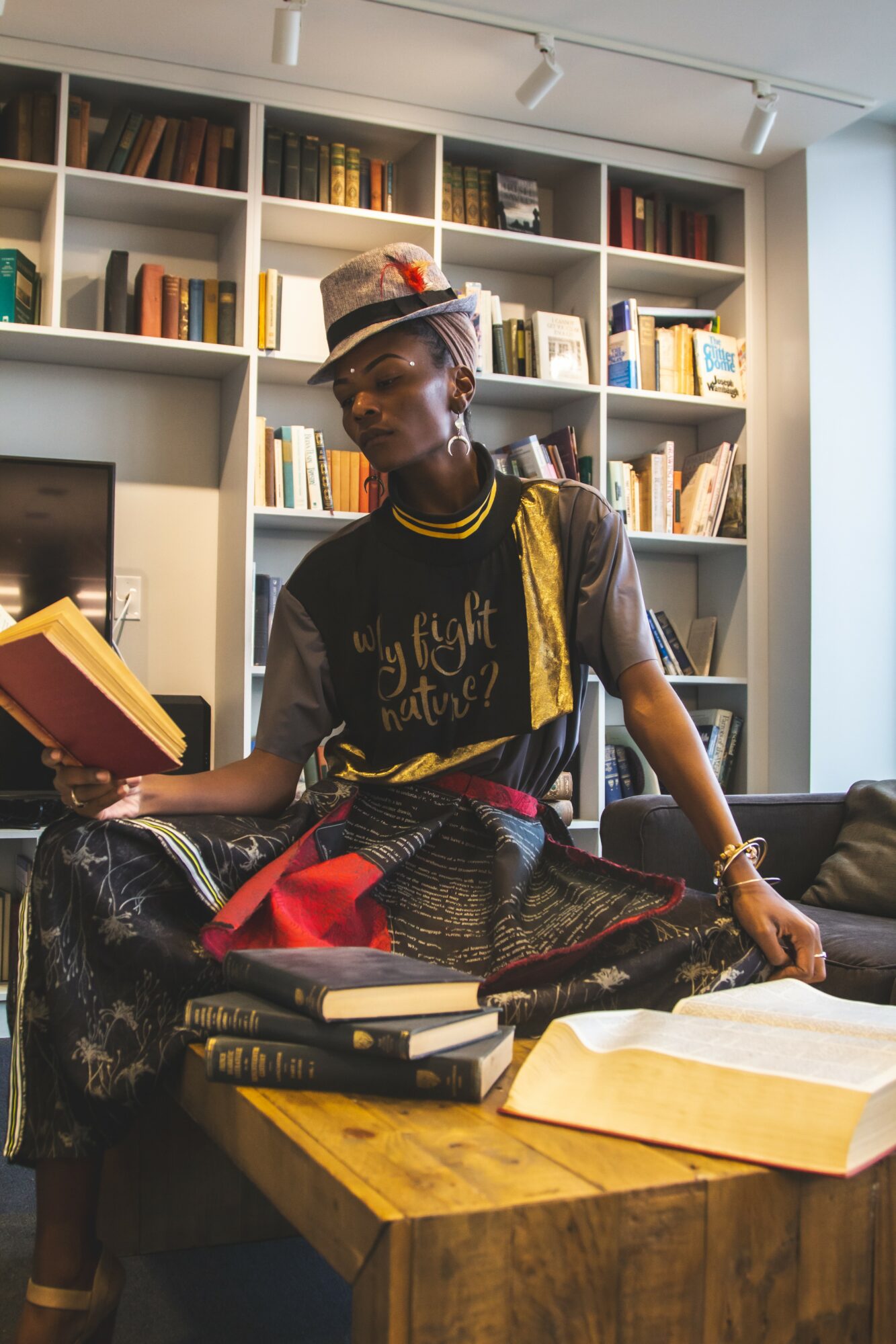 Person sitting on a table reading a book, surrounded by books and a bookshelf in the background.