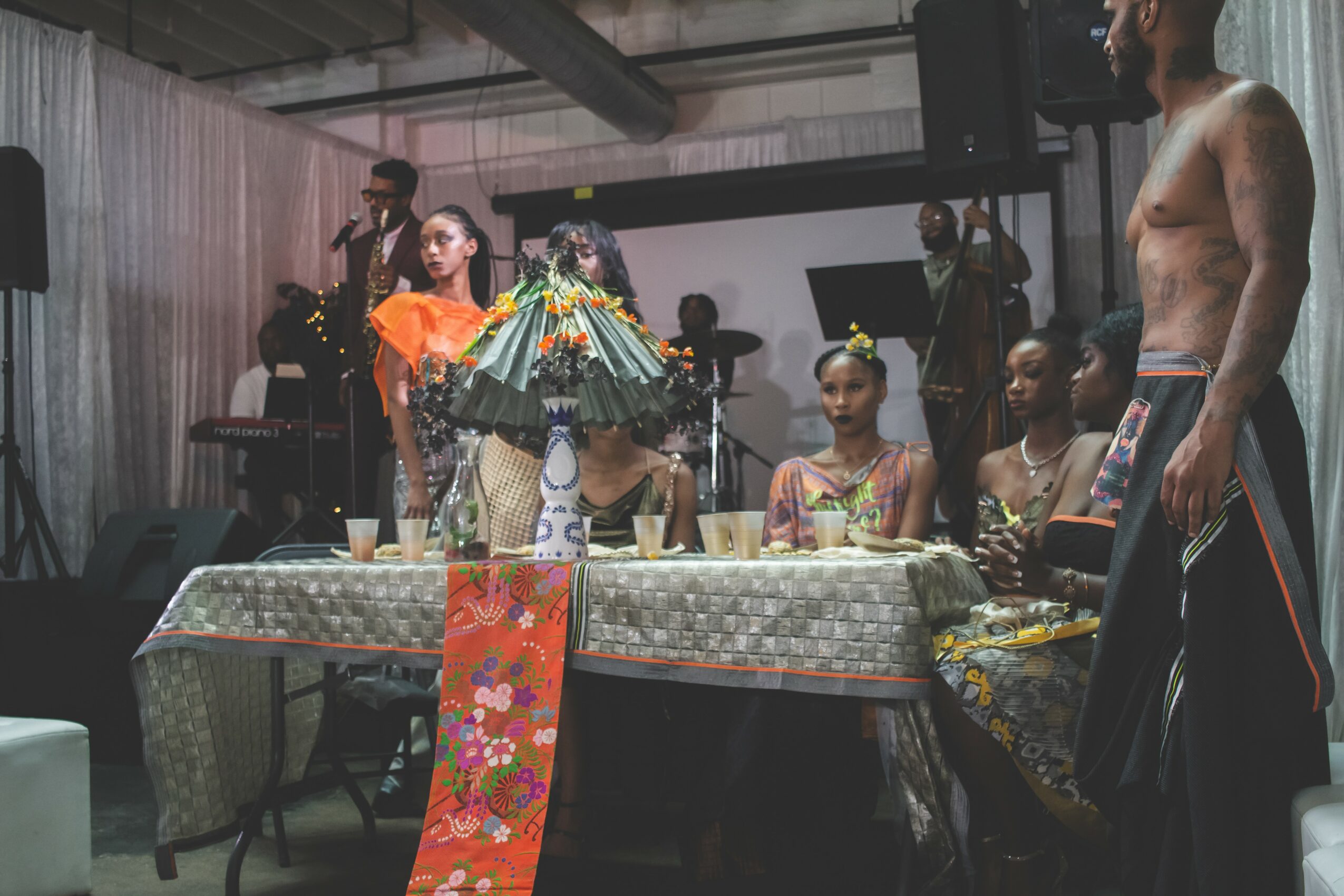 People in traditional costumes around a decorated table with a colorful umbrella, in an indoor setting with musicians in the background.