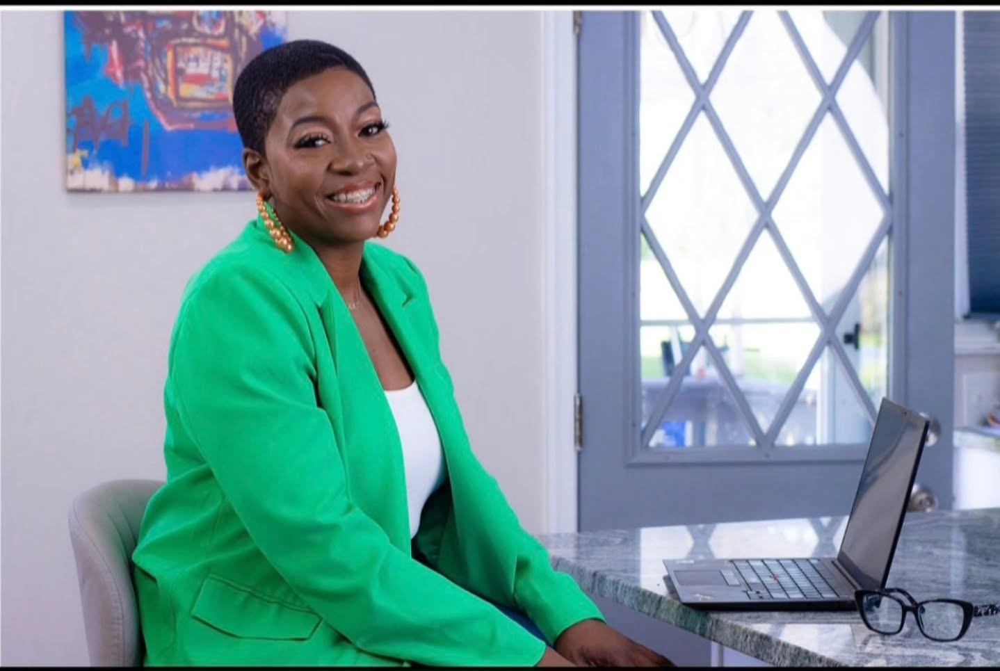 Smiling woman in green blazer sitting at desk with laptop and glasses, in a bright room with window.