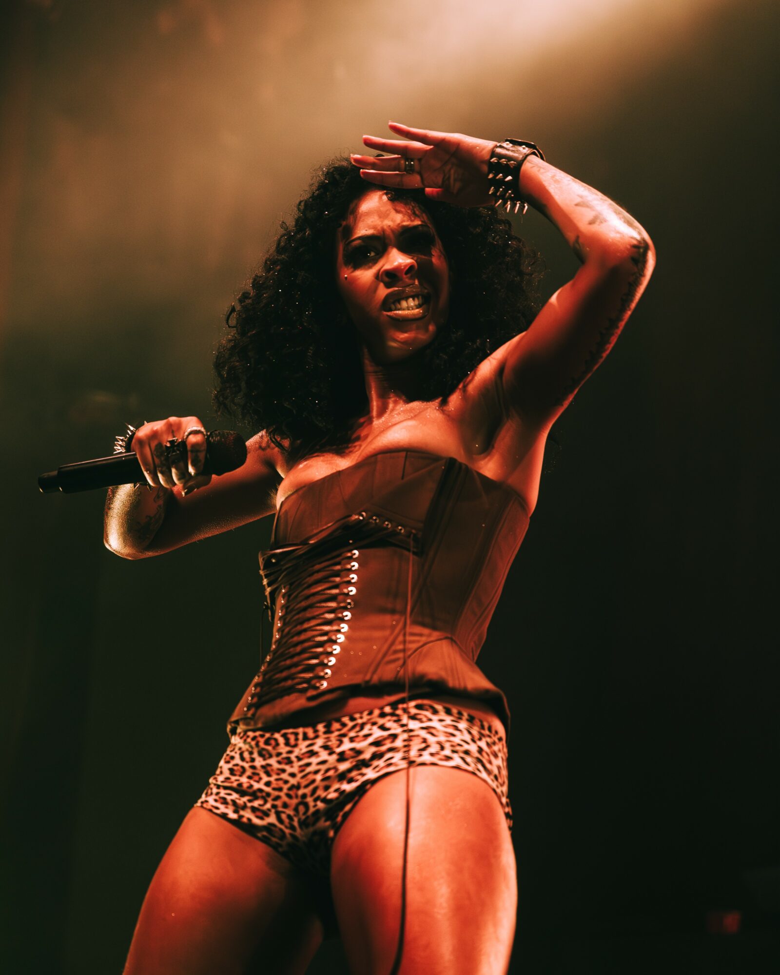 Performer on stage with curly hair, wearing a corset and leopard print shorts, holding a microphone, with a hand raised to her forehead.