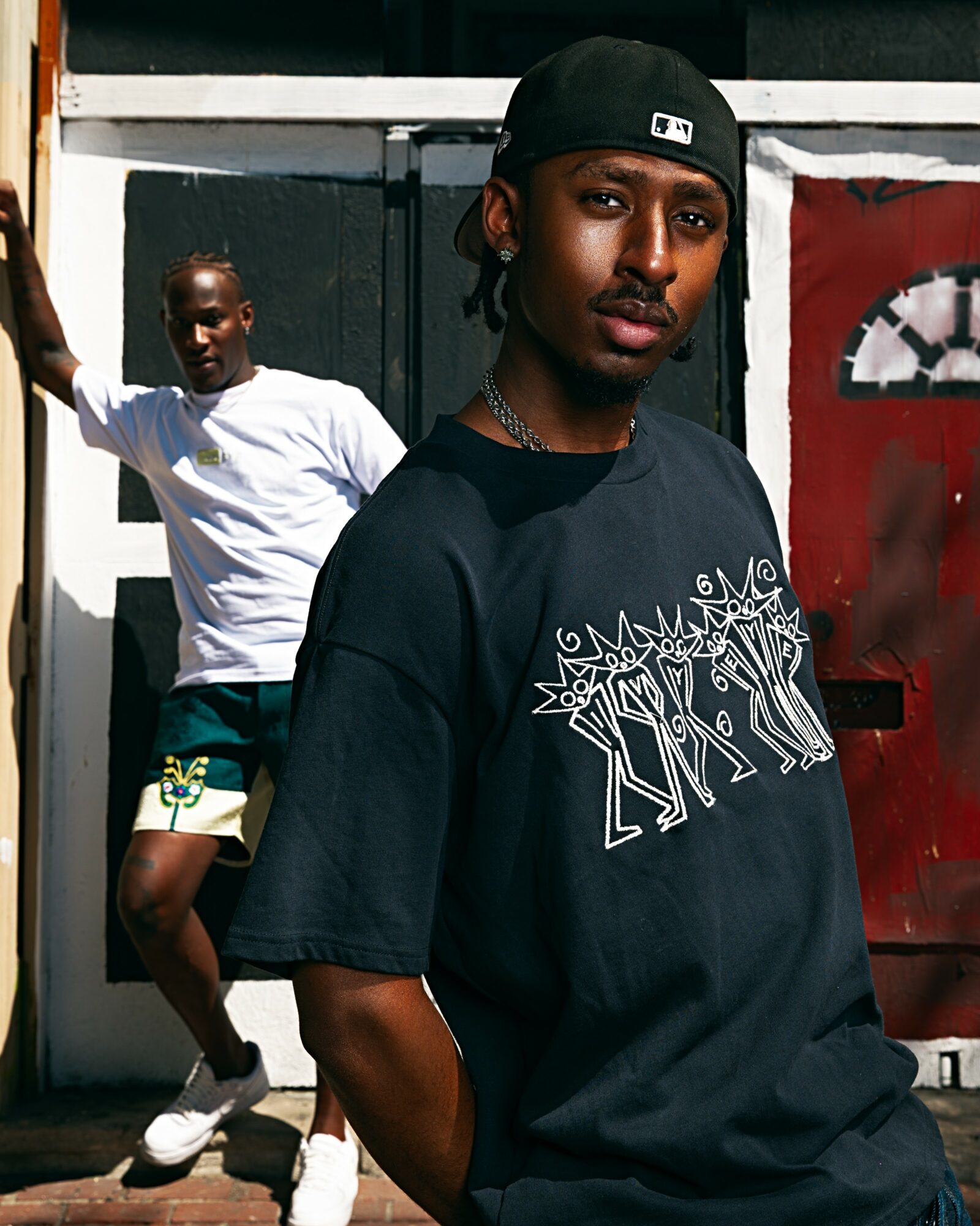 Two young men outdoors, one in the foreground wearing a black cap and T-shirt, the other in the background in a white T-shirt and shorts.