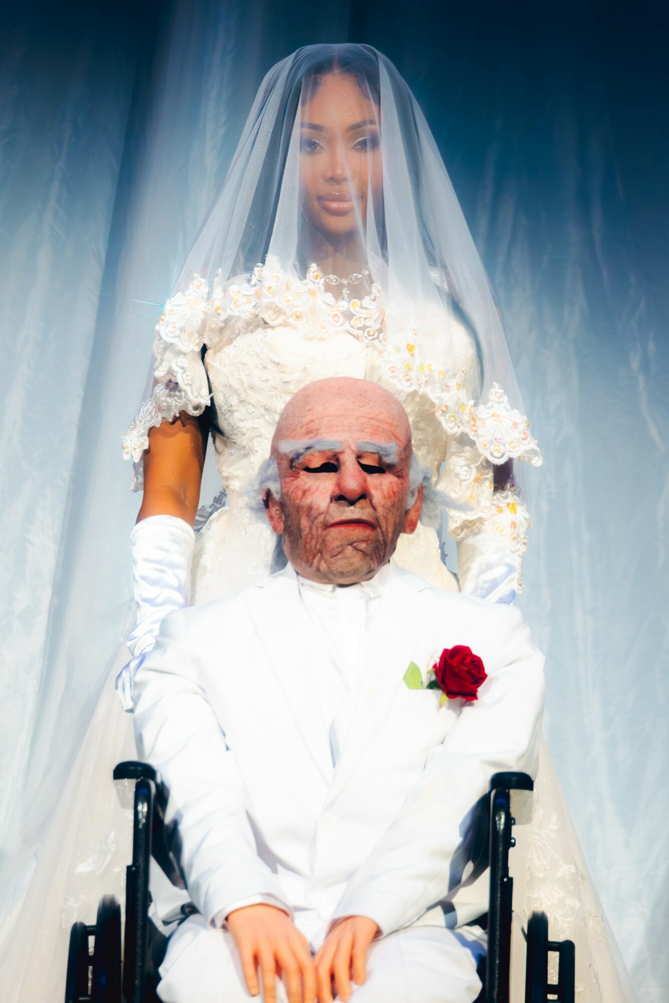A young woman in a wedding dress stands behind an elderly man in a wheelchair, with a veil covering her head.