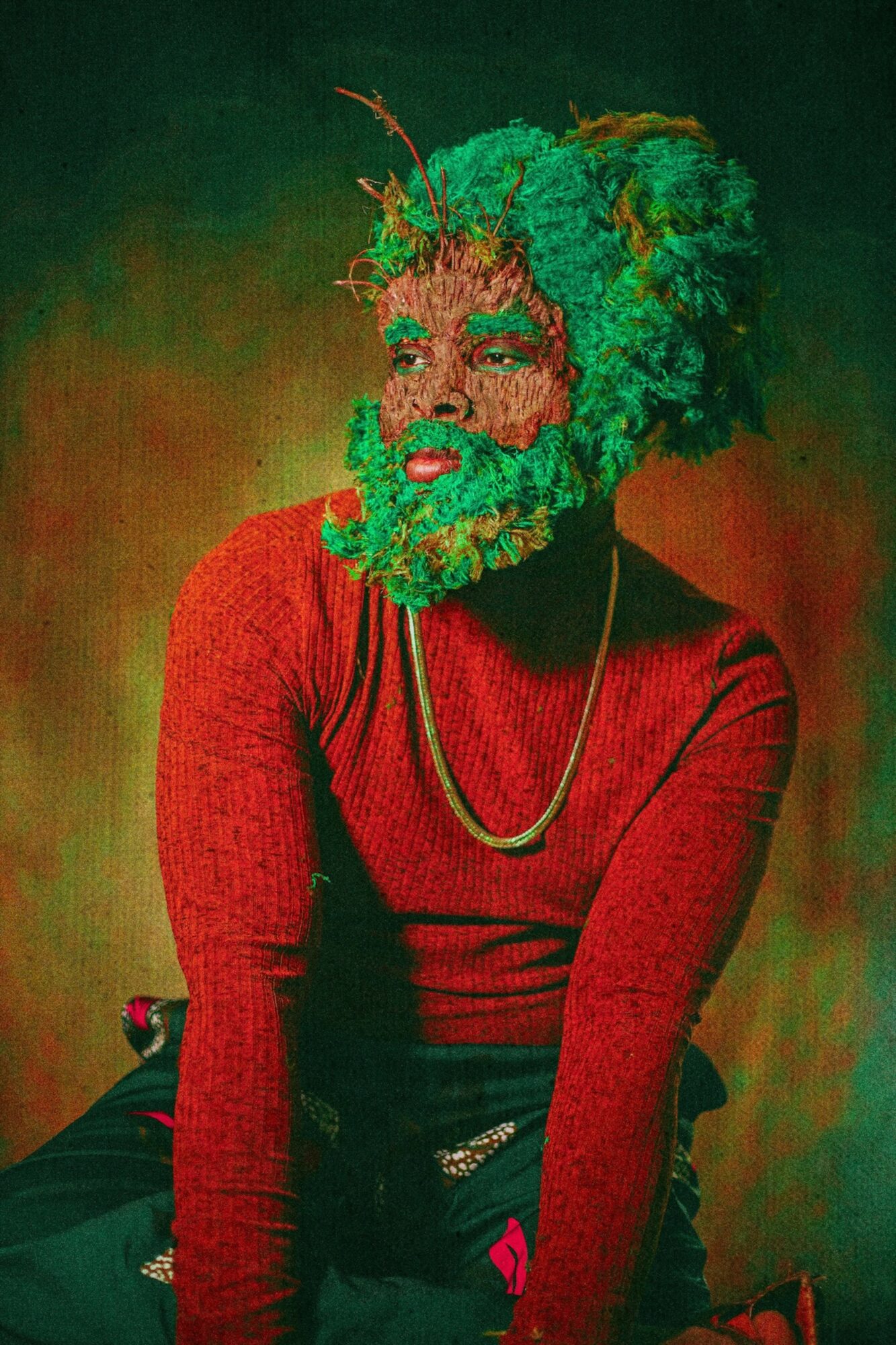 Person with green hair and beard, wearing a red top and gold necklace, sitting against a colorful background.
