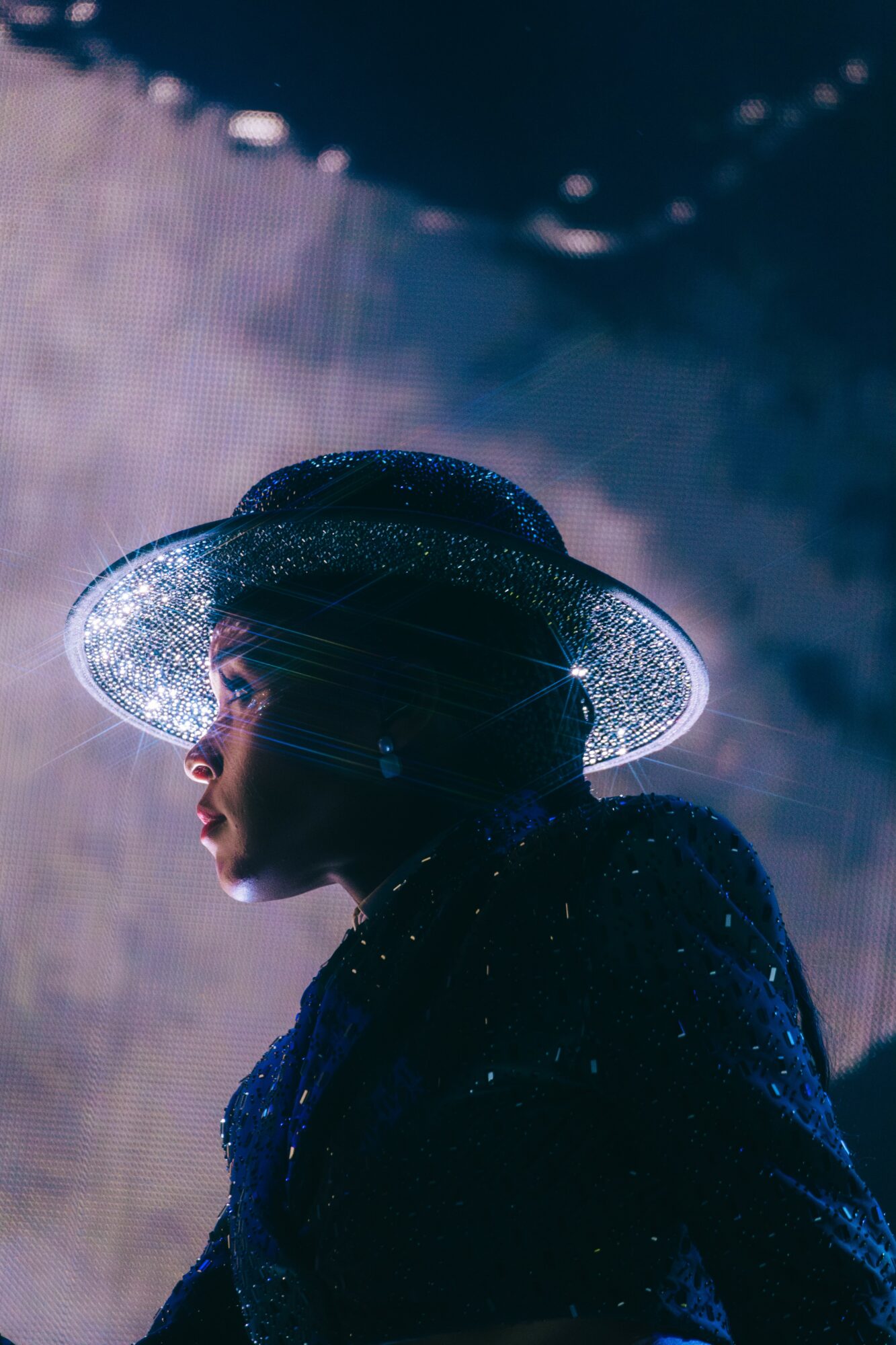 Person wearing a wide-brimmed, glittering hat, facing left, with a dark, sparkly outfit, against a dark background.