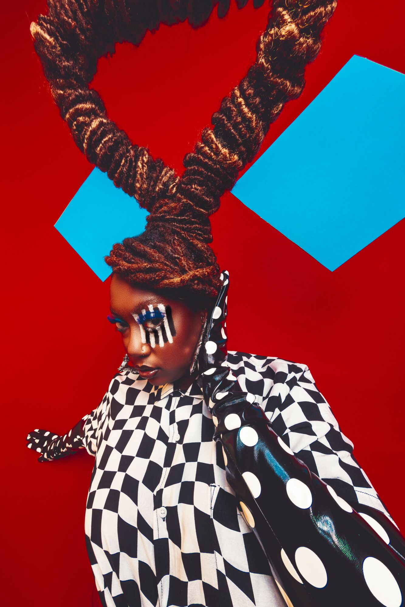 Person with large braided hair, wearing a checkered outfit, against a red background with blue paper, looking down.
