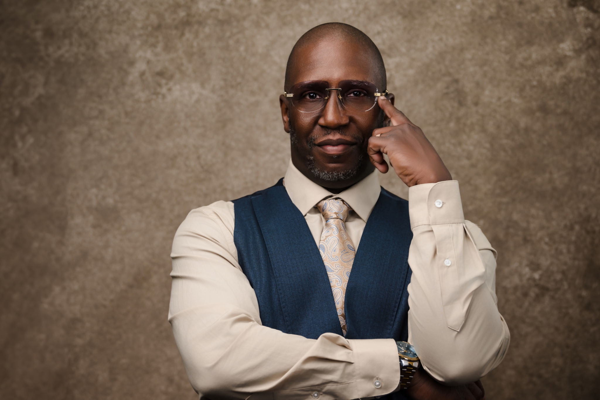 Man with glasses in a white shirt, blue vest, and tie, posing with hand near face against a textured background.