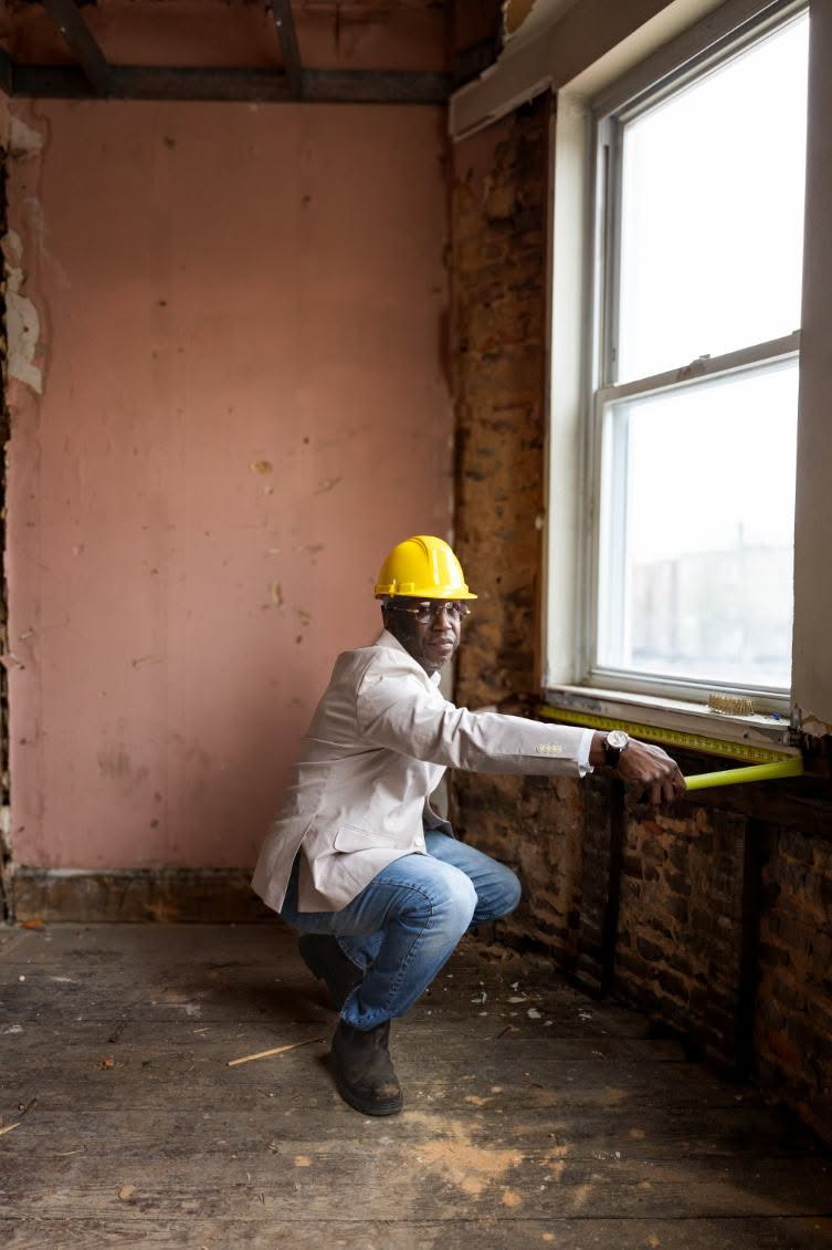 Person wearing a yellow hard hat squatting by a window, measuring with a yellow level inside a partially renovated room.