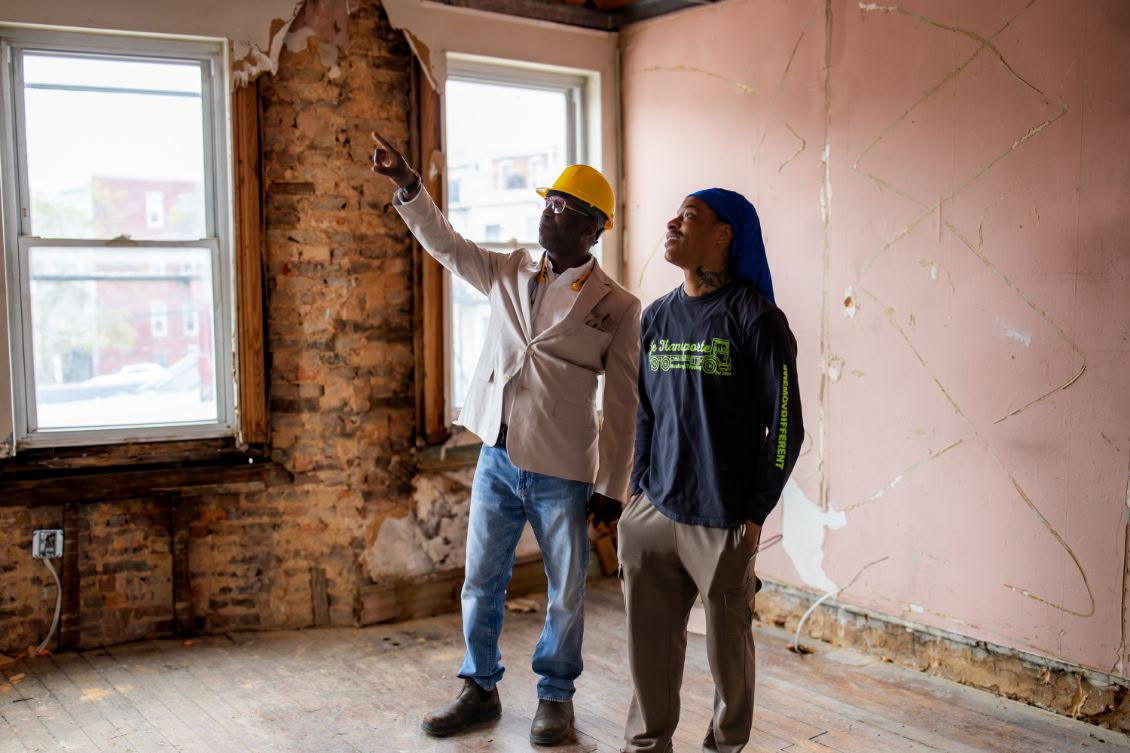 Two people in a room with exposed brick and peeling pink paint, one pointing at the wall, windows in the background.