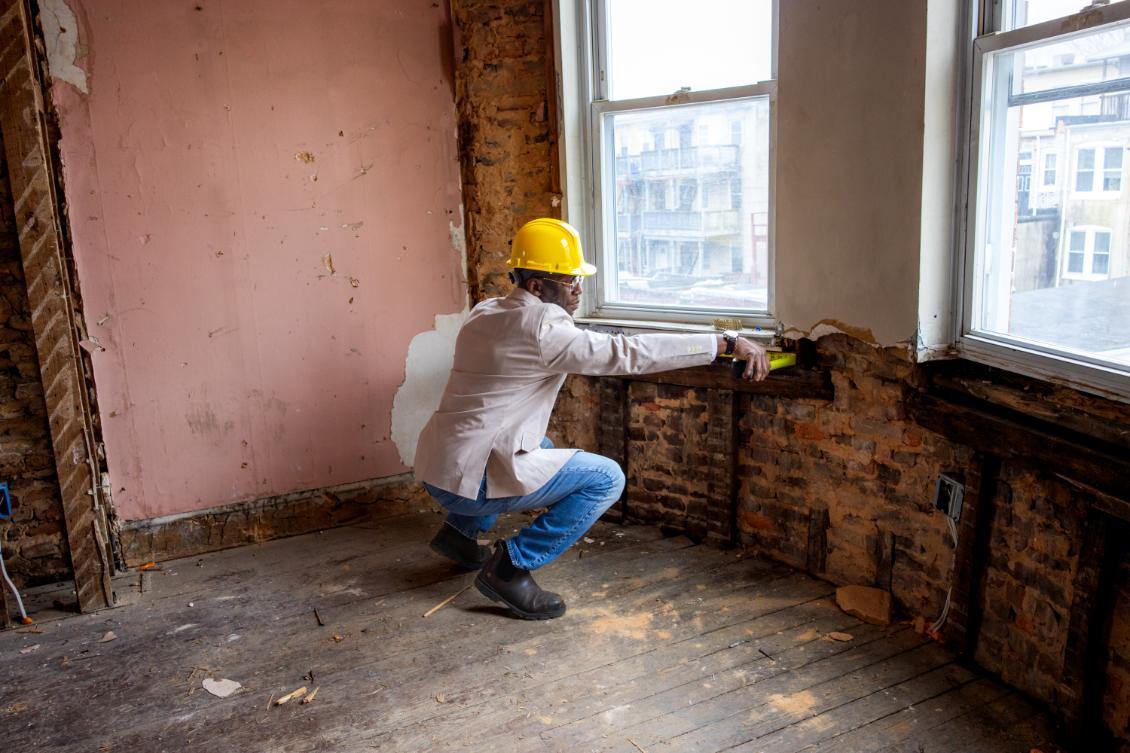 Person wearing a yellow helmet cleaning a window in a room with exposed brick and peeling paint, sunlight coming through windows.