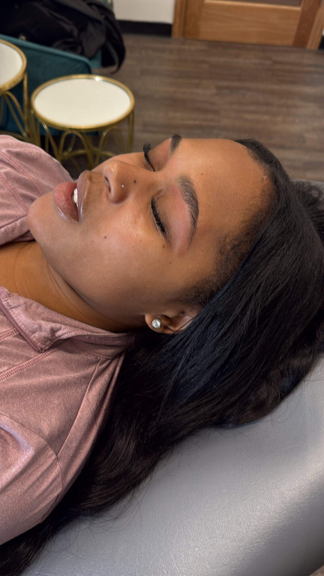 Woman with closed eyes lying down, dark hair, pink top, in a room with chairs and wooden floor.