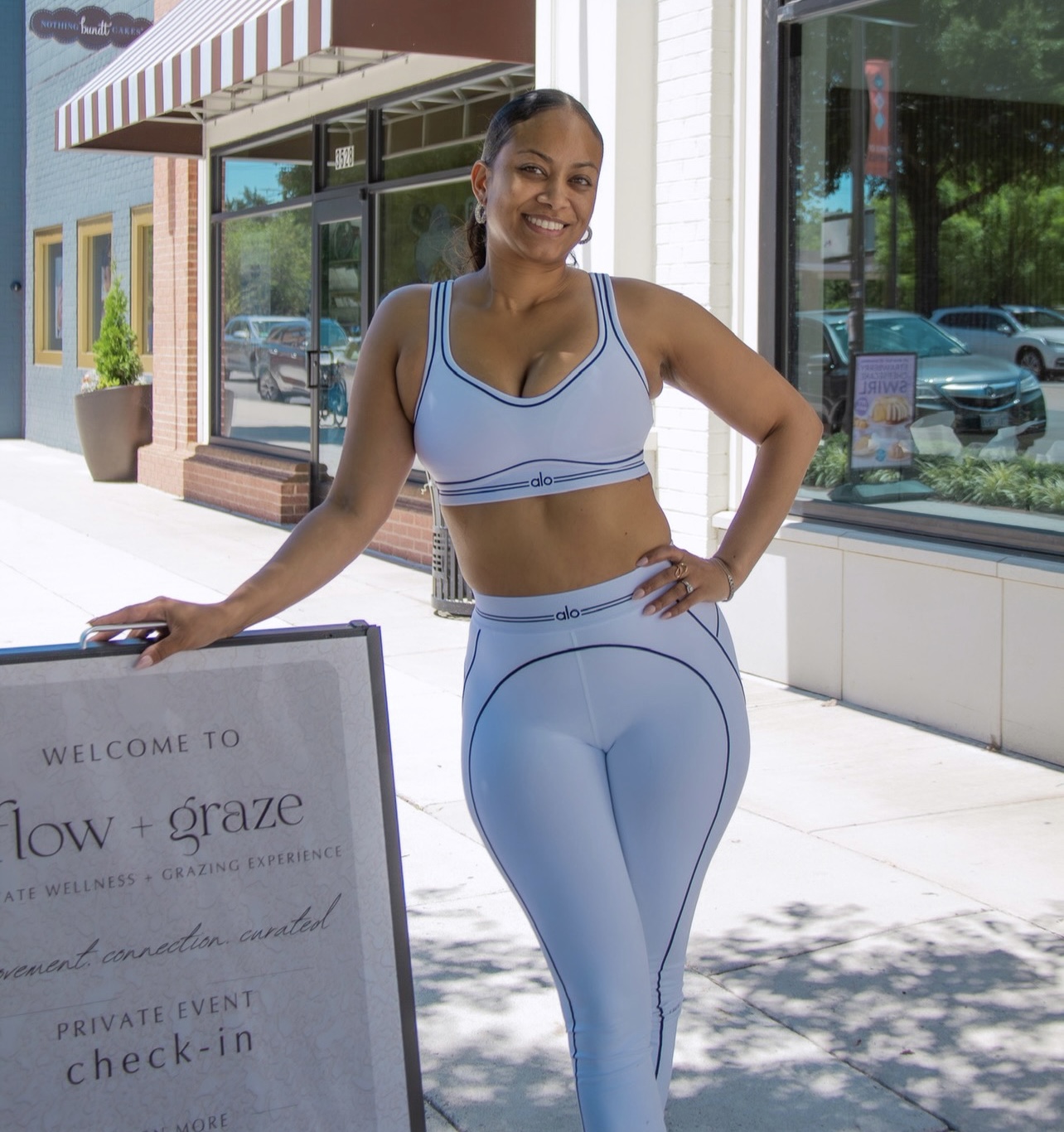 Smiling woman in workout clothes standing outside a storefront with large windows and a sidewalk.