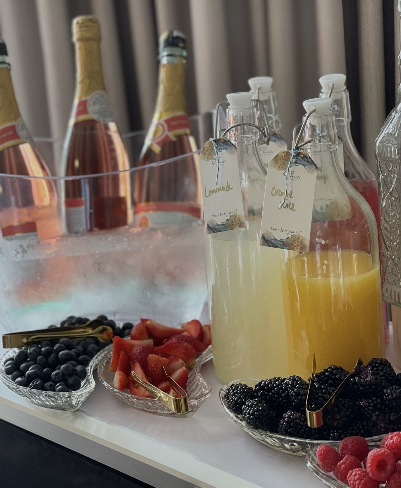 Bottles of sparkling wine, juice, and water with fruit bowls and glasses on a table, curtains in the background.
