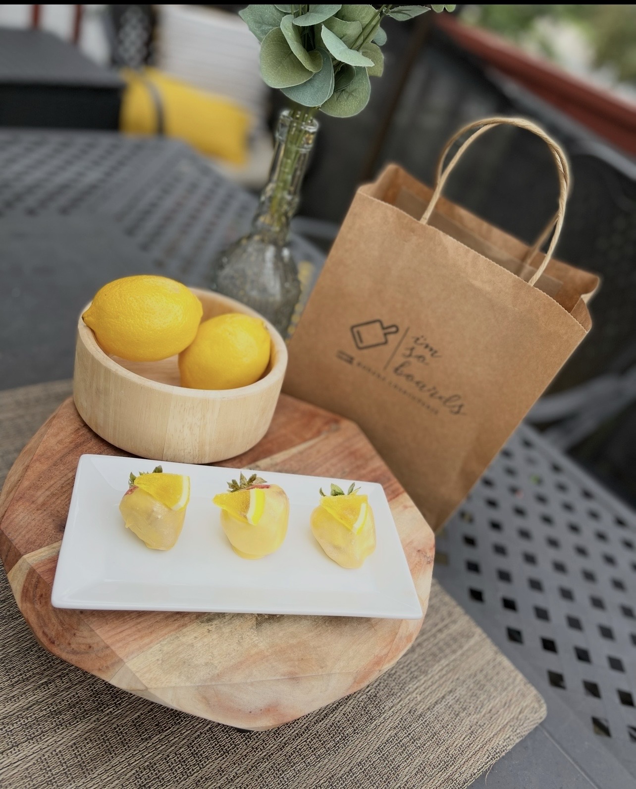 Lemon and lemon desserts on a white plate, a paper bag, and a wooden bowl with lemons on a table, with a plant in the background.