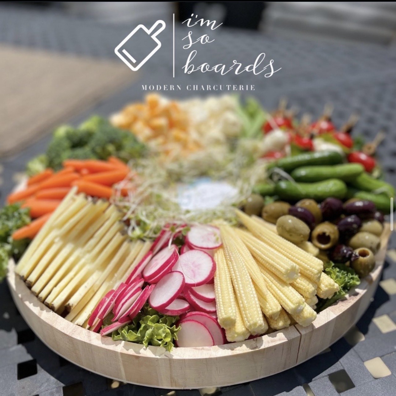 Assorted fresh vegetables and cheeses arranged on a round wooden platter, with a logo and text overlay at the top.