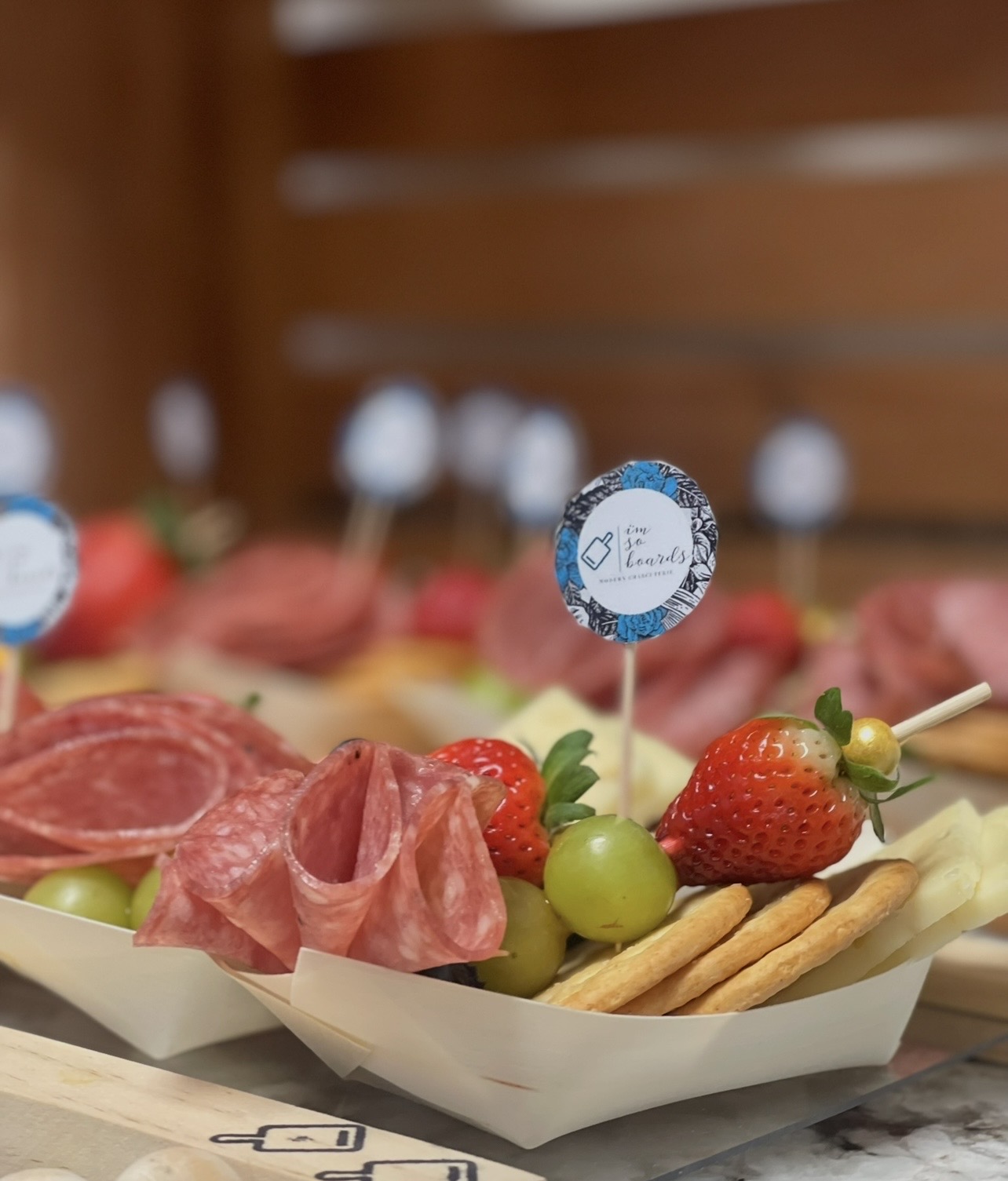 Tray with strawberries, green grapes, salami, crackers, and a small sign, on a wooden surface with a blurred background.