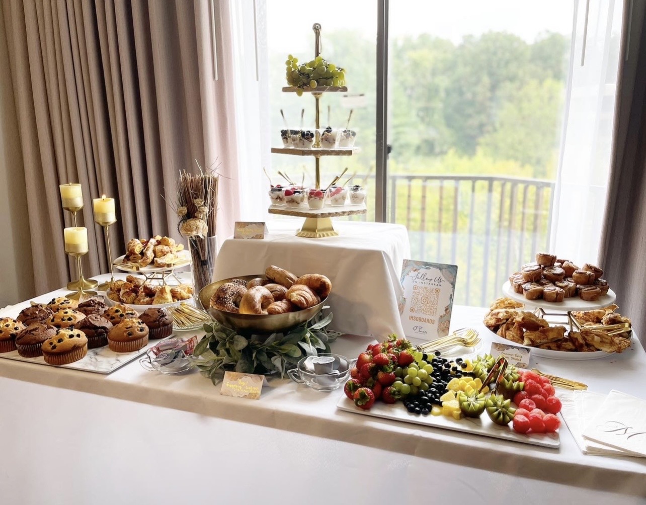Dessert table with cupcakes, bread rolls, fruit, and a three-tiered stand, set near a window with a balcony view.