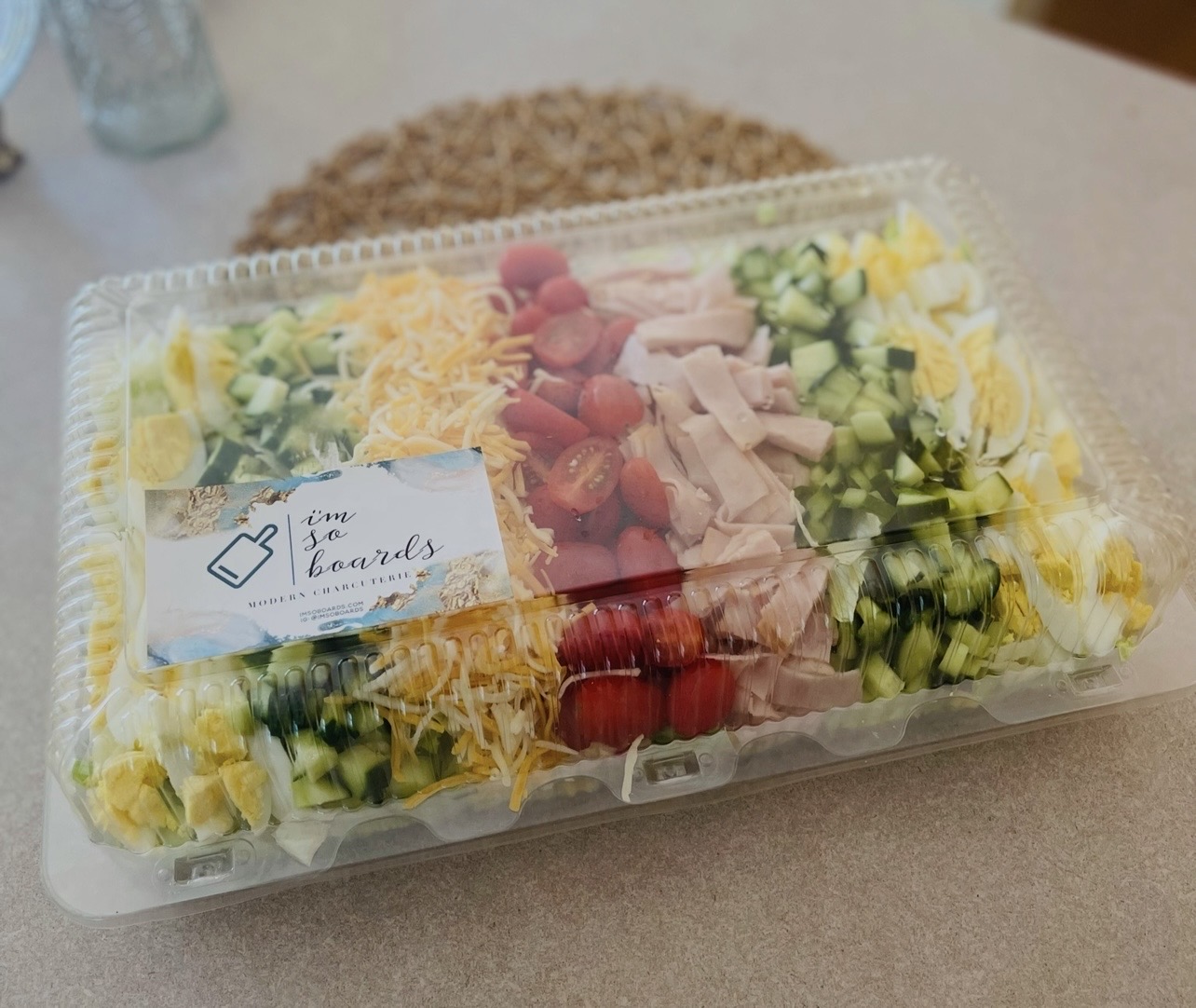 Clear plastic container with a salad containing cherry tomatoes, shredded cheese, sliced chicken, and chopped cucumbers, on a beige surface.