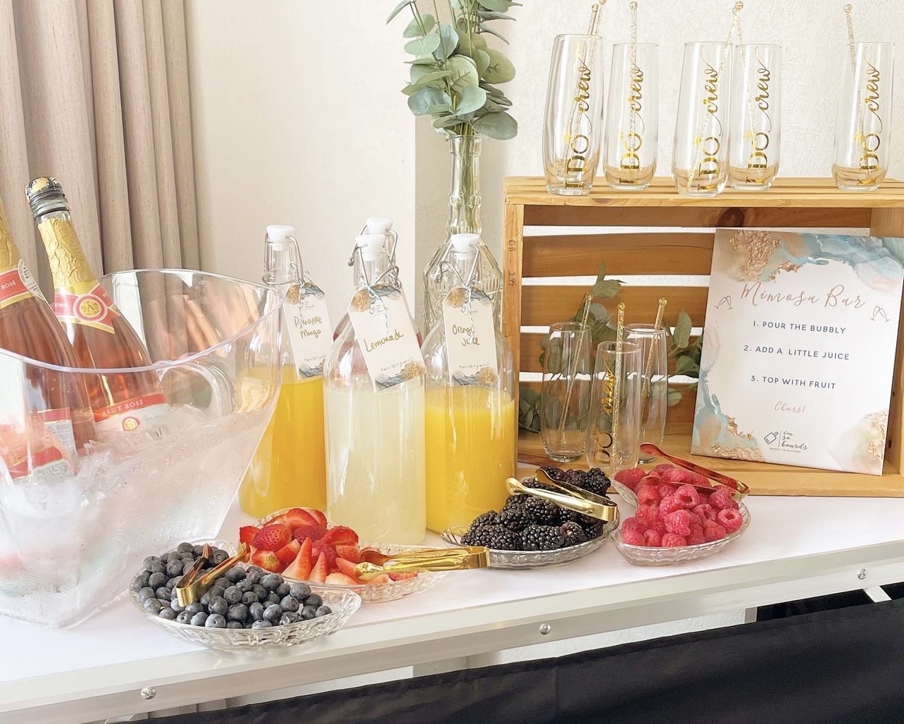 Beverage station with bottles, glasses, and fruit garnishes on a table, with a framed sign and decorative items in the background.