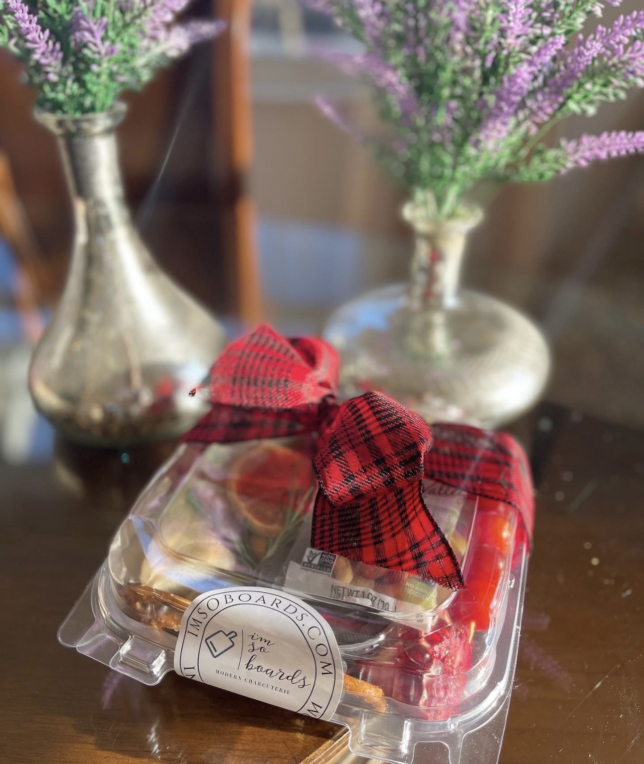 Clear plastic container with red and white checkered ribbon, placed on a wooden surface with purple flowers in vases in background.