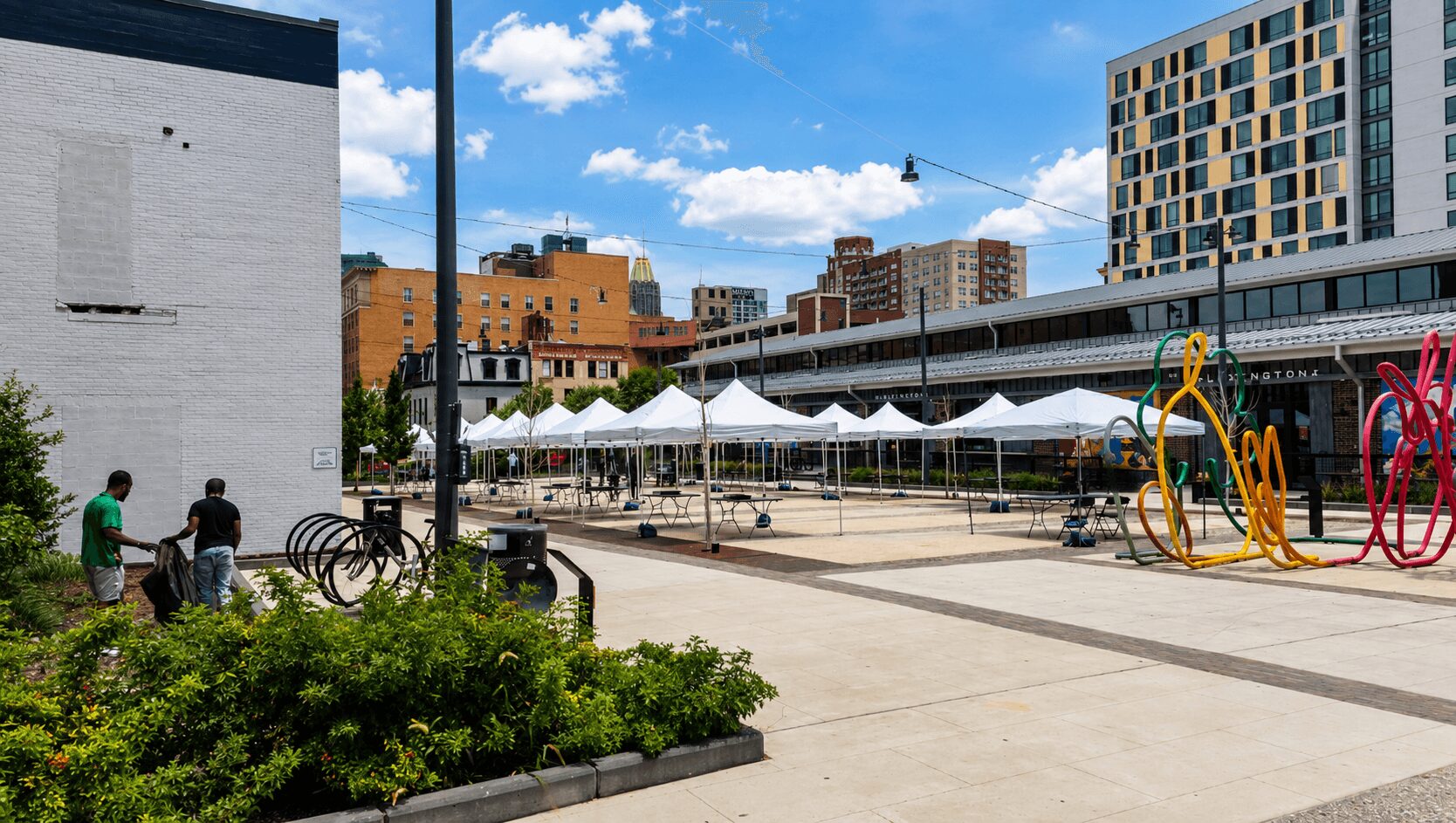 Cityscape with tents, modern buildings, sculptures, and people walking in an outdoor plaza.
