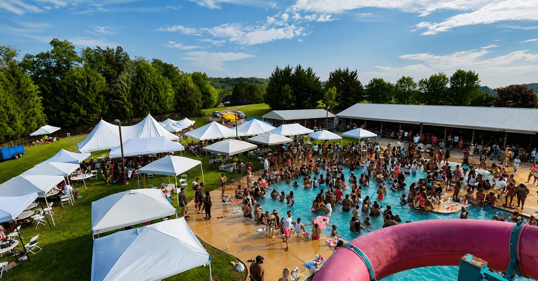 Crowd of people in a swimming pool with white tents and trees in the background, sunny day.