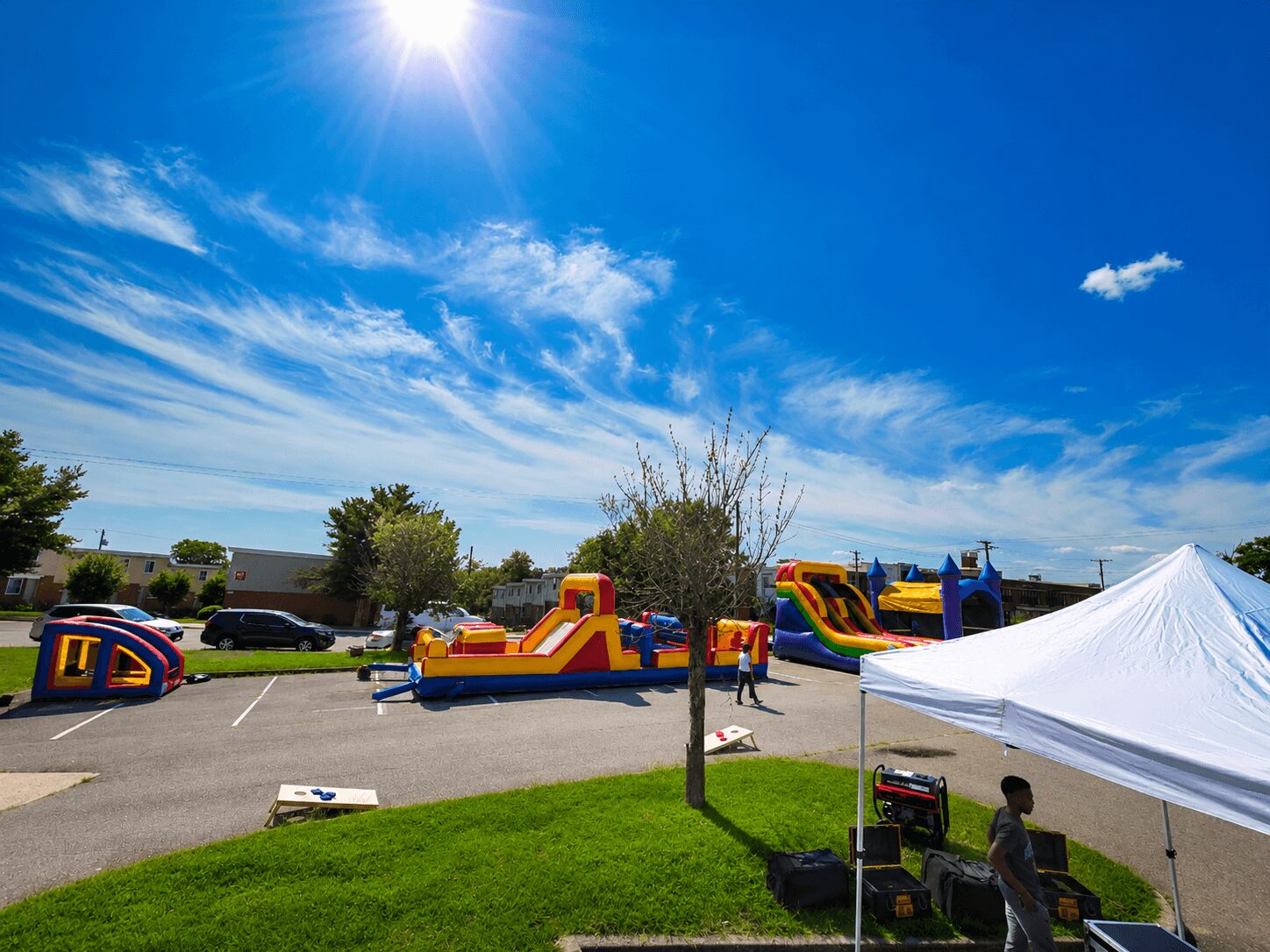 Inflatable bounce houses and slides set up in a parking lot with a grassy area, under a bright blue sky with clouds.