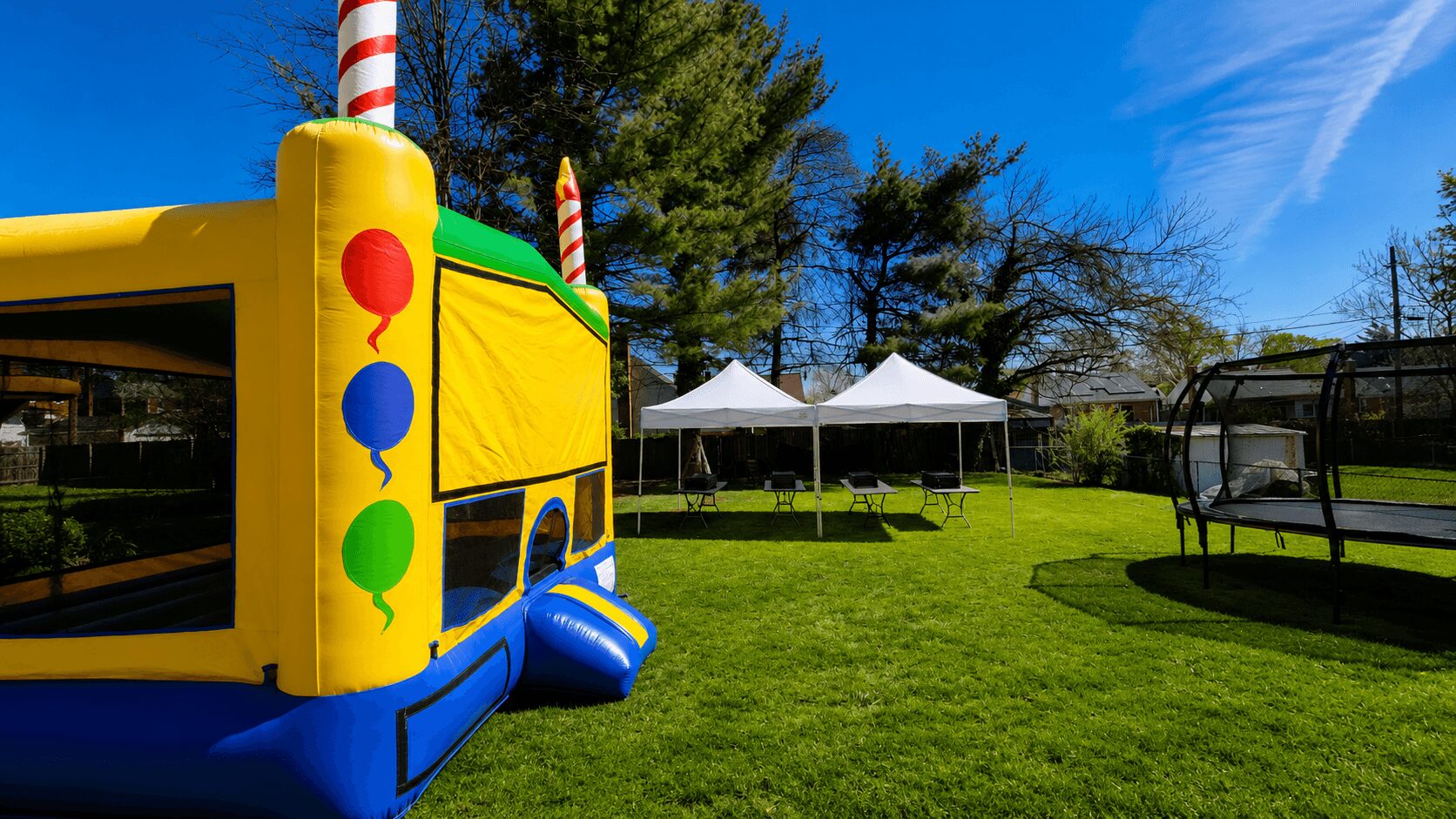 Play area with inflatable bounce house, two white tents, and outdoor seating on a grassy field under a blue sky.