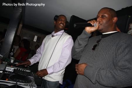 Two men are performing on stage, one singing into a microphone and the other smiling at a DJ setup.