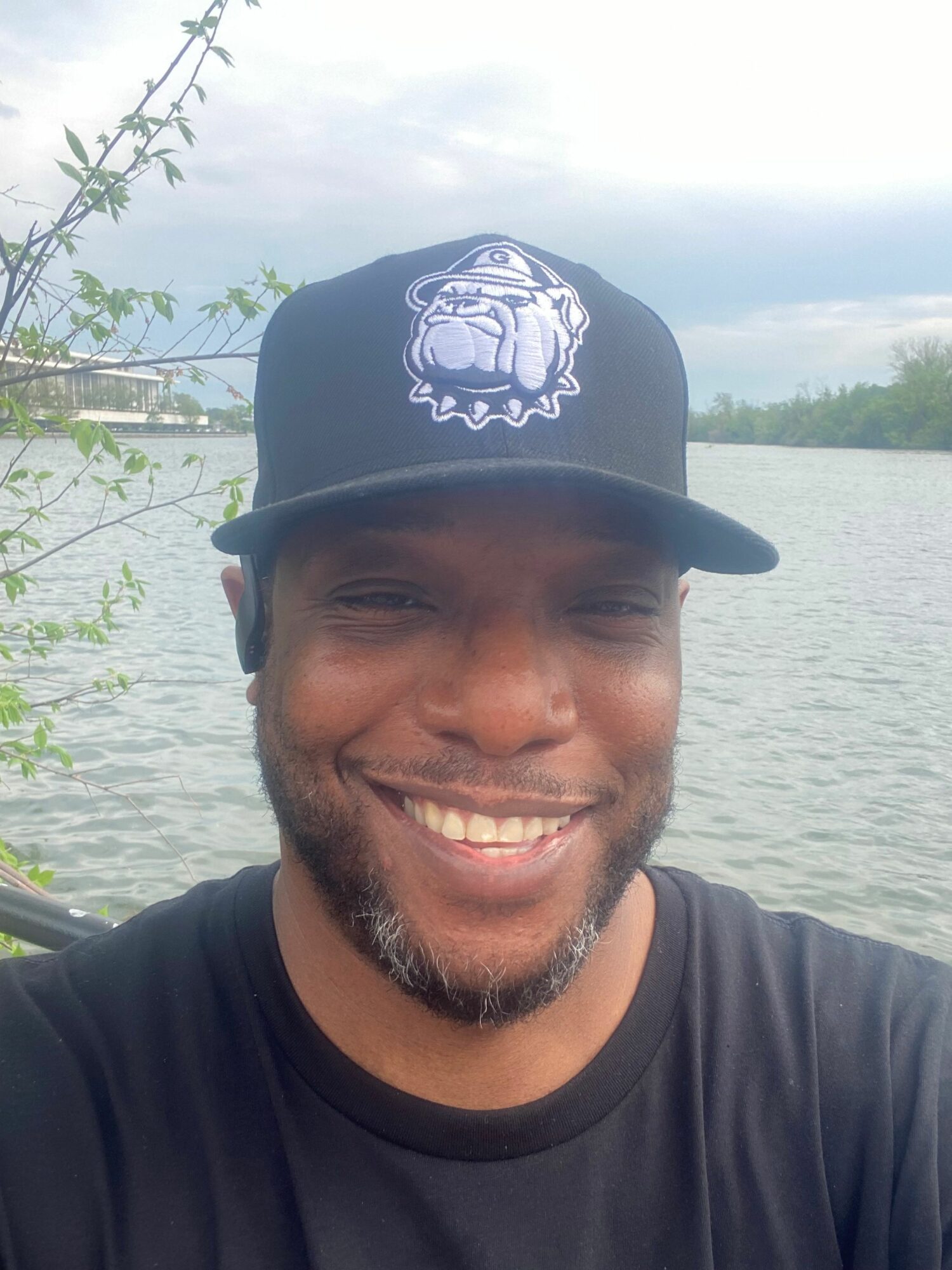Smiling man wearing a black cap with a lion logo, outdoors near water, with trees and sky in background.