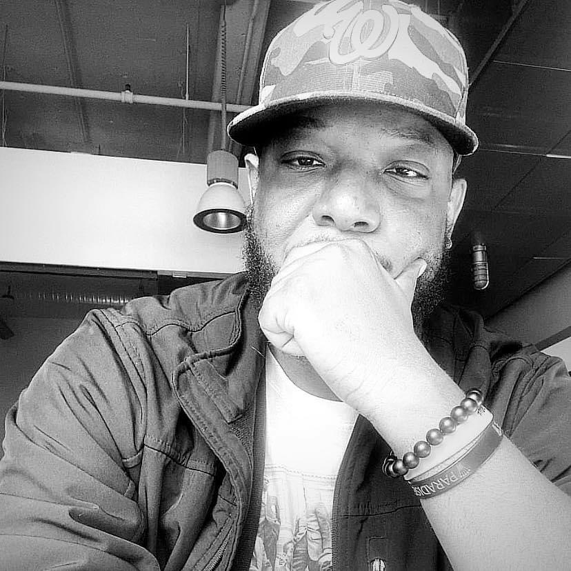 Young man wearing a cap and jacket, resting his chin on his hand, in an indoor setting.