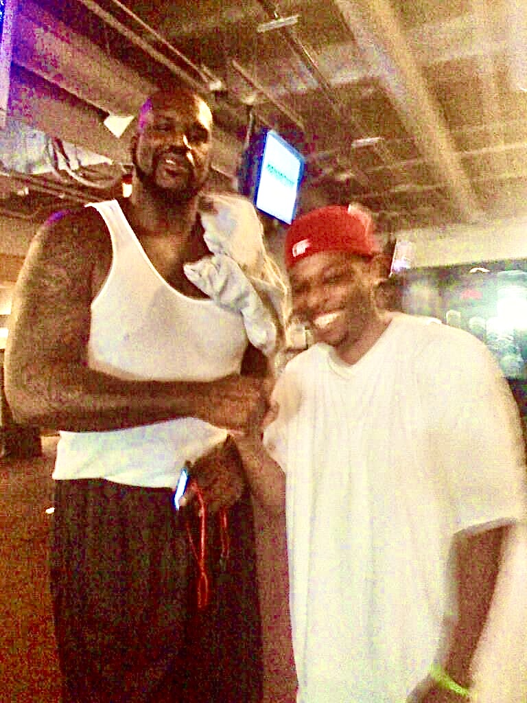 Two people smiling, one wearing a white tank top and the other in a white shirt and red cap, in a dimly lit indoor setting.