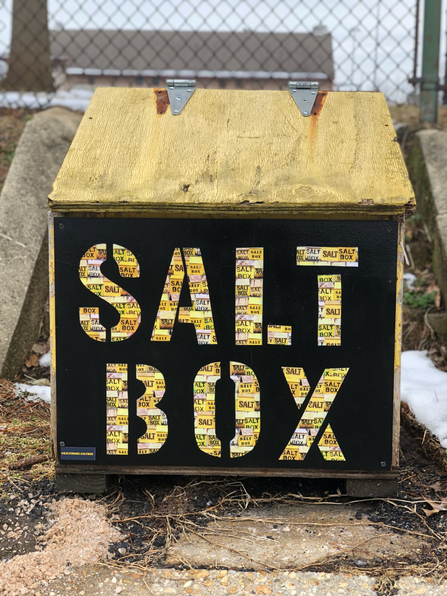 A black salt box with a yellow lid, labeled 'SALT BOX' in large letters, outdoors near a chain-link fence.