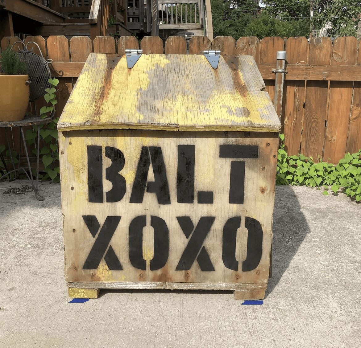 Wooden box with the words 'BALT XOXO' painted on it, placed outdoors on a concrete surface with a wooden fence in the background.