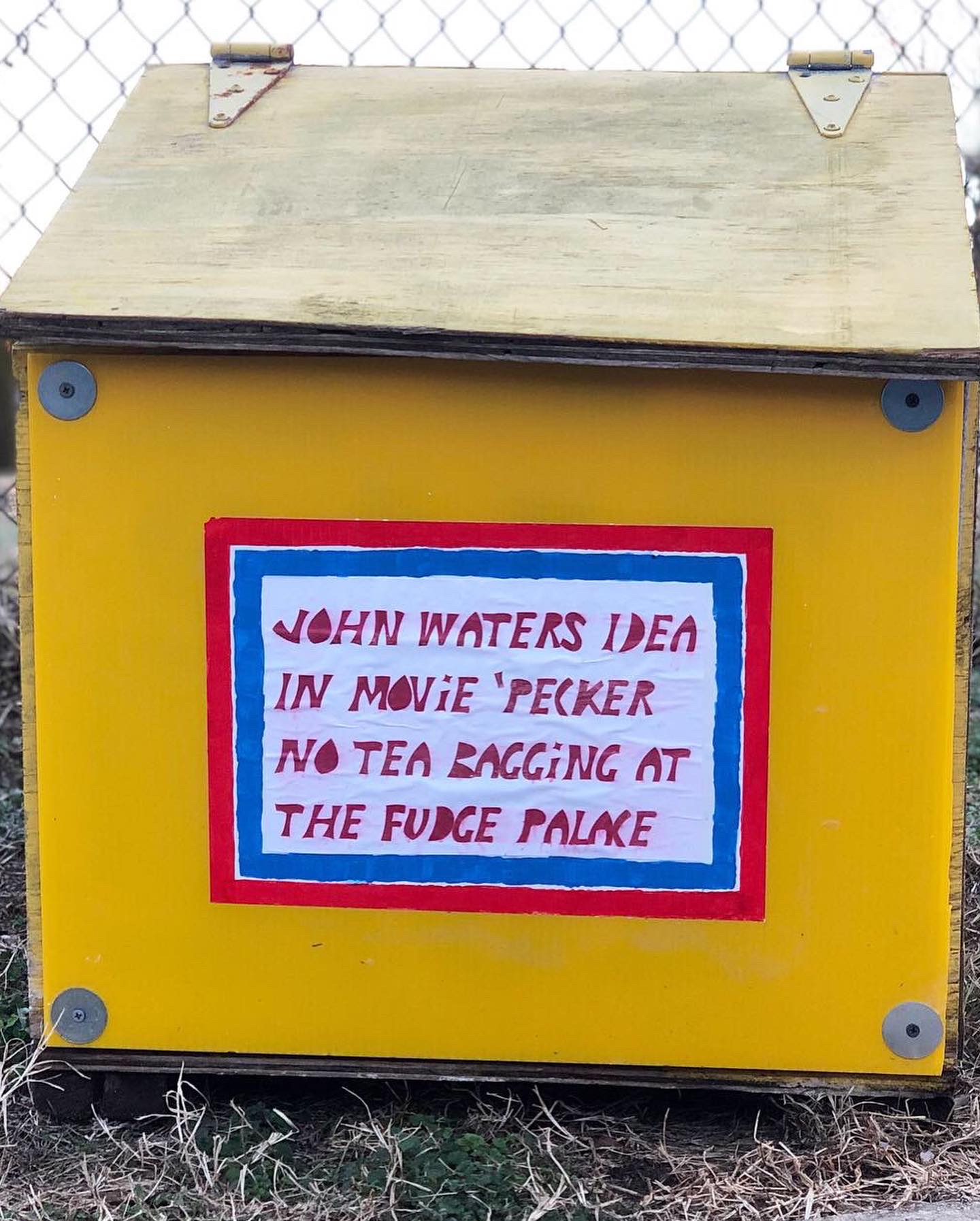 Yellow box with a handwritten note on it, attached to a fence, with grass at the bottom.