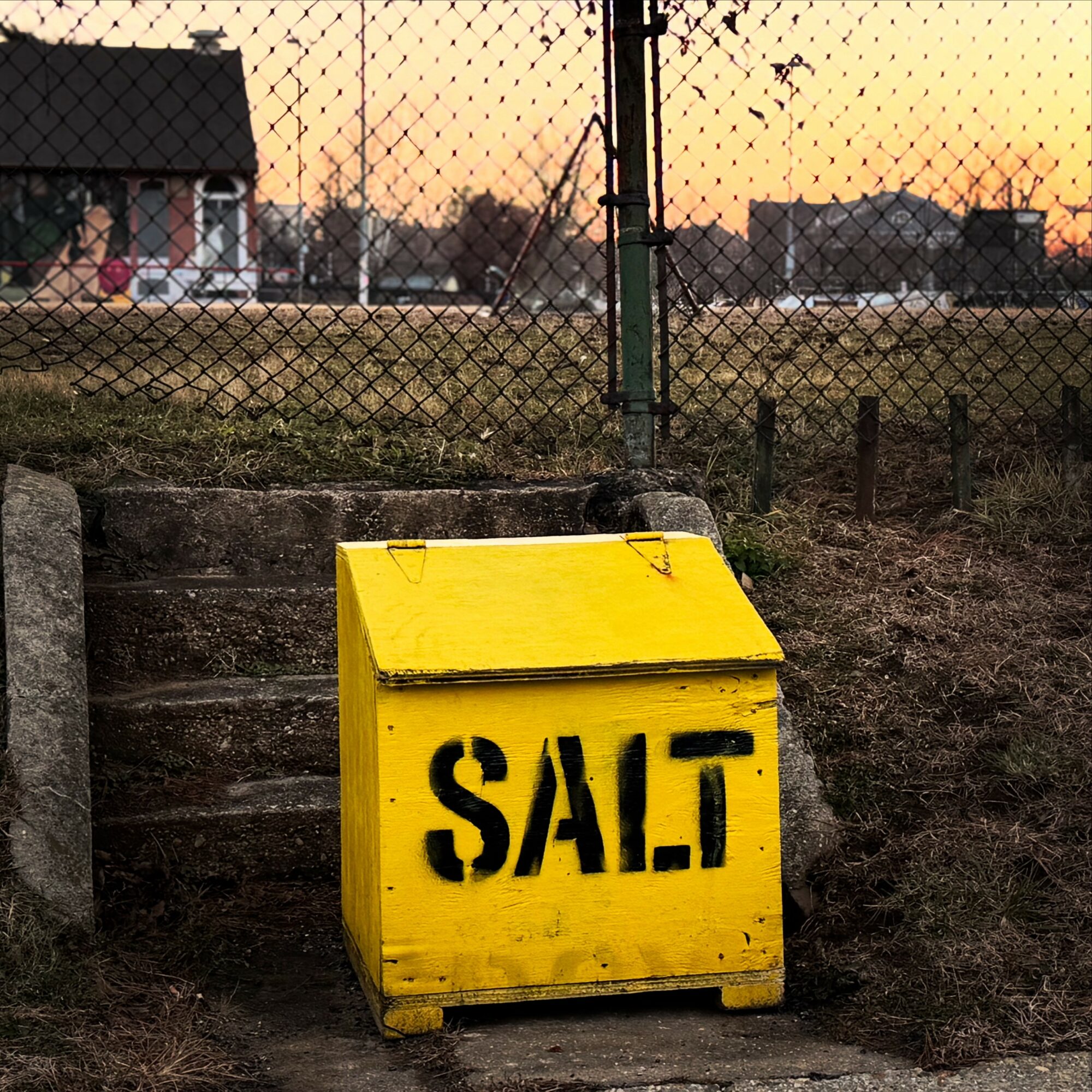 Yellow salt storage box with black text, outdoors near a fence and steps, sunset sky in background.