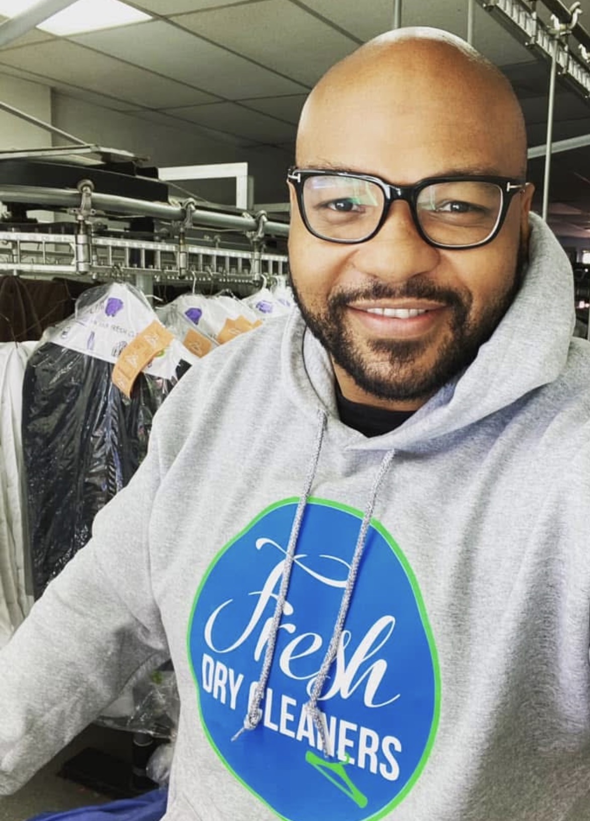 Man with glasses and a beard smiling, wearing a gray hoodie with a blue logo, in a laundry or dry cleaning store.
