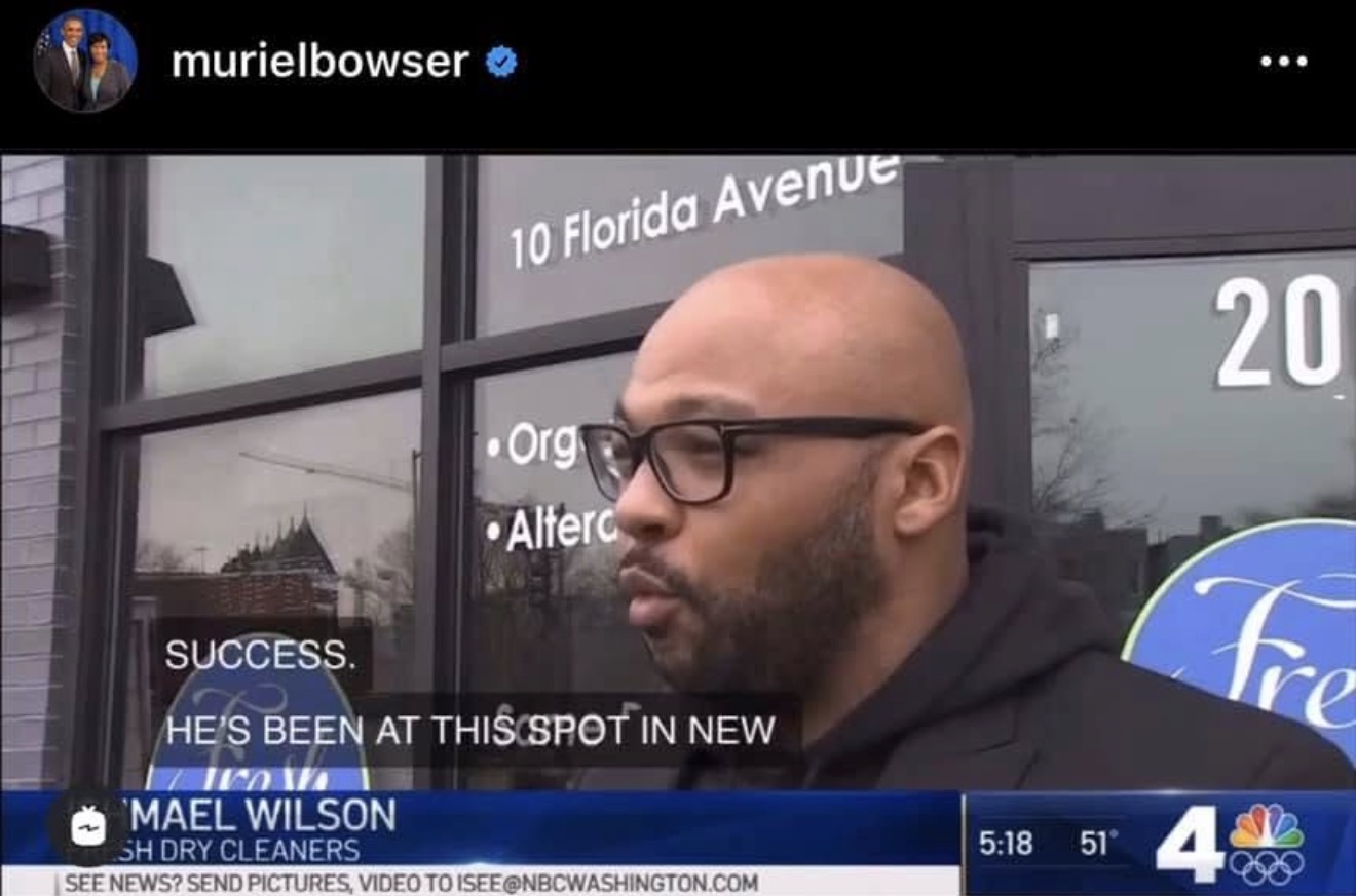 Man with glasses and a beard in front of a building with signs, speaking to the camera, with a news overlay.
