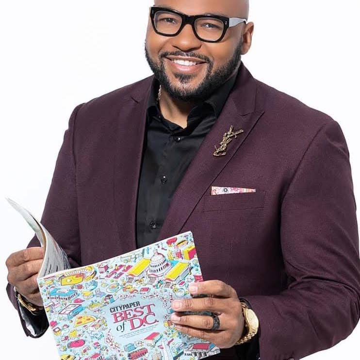 Man with glasses, beard, and dark skin, smiling, holding a colorful magazine, wearing a maroon blazer and black shirt.