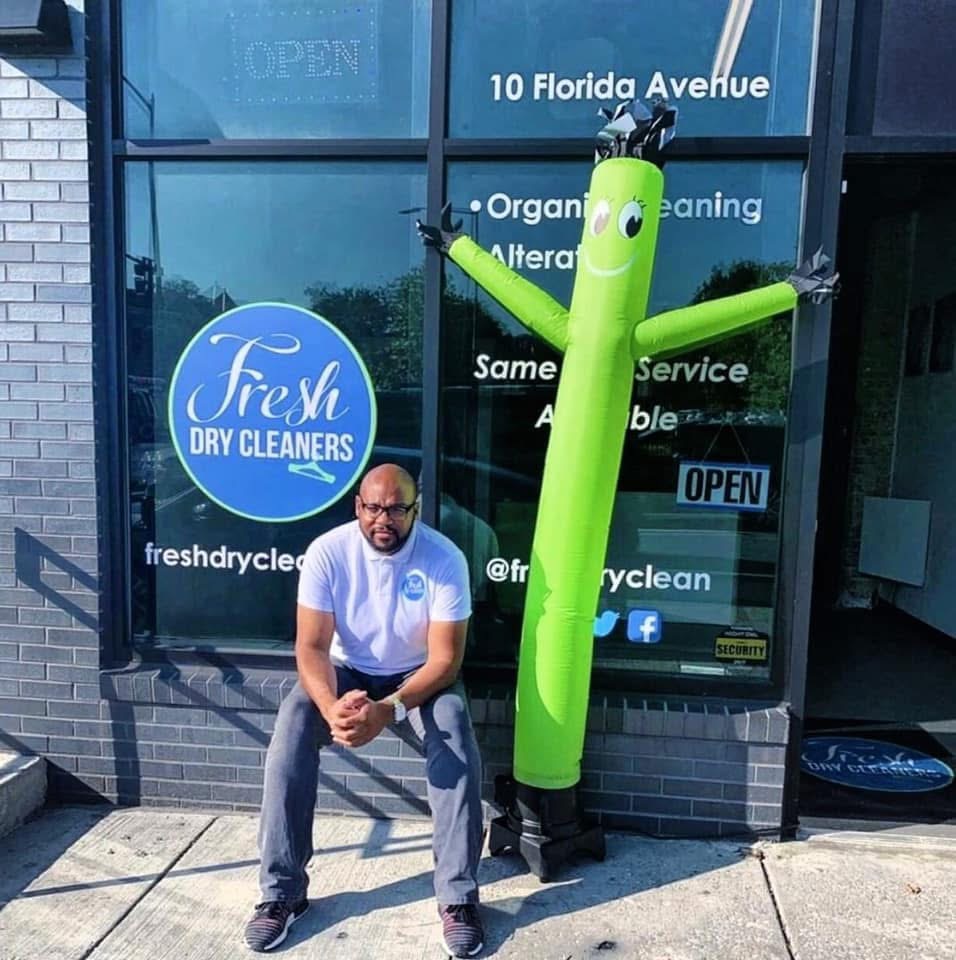 Person crouching next to a green inflatable tube man outside a dry cleaner store with glass windows and signage.