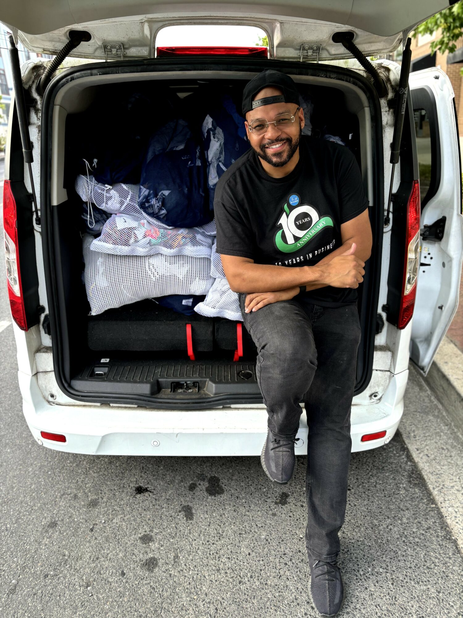 Man smiling, sitting on the edge of an open van trunk filled with bags and boxes, on a street.