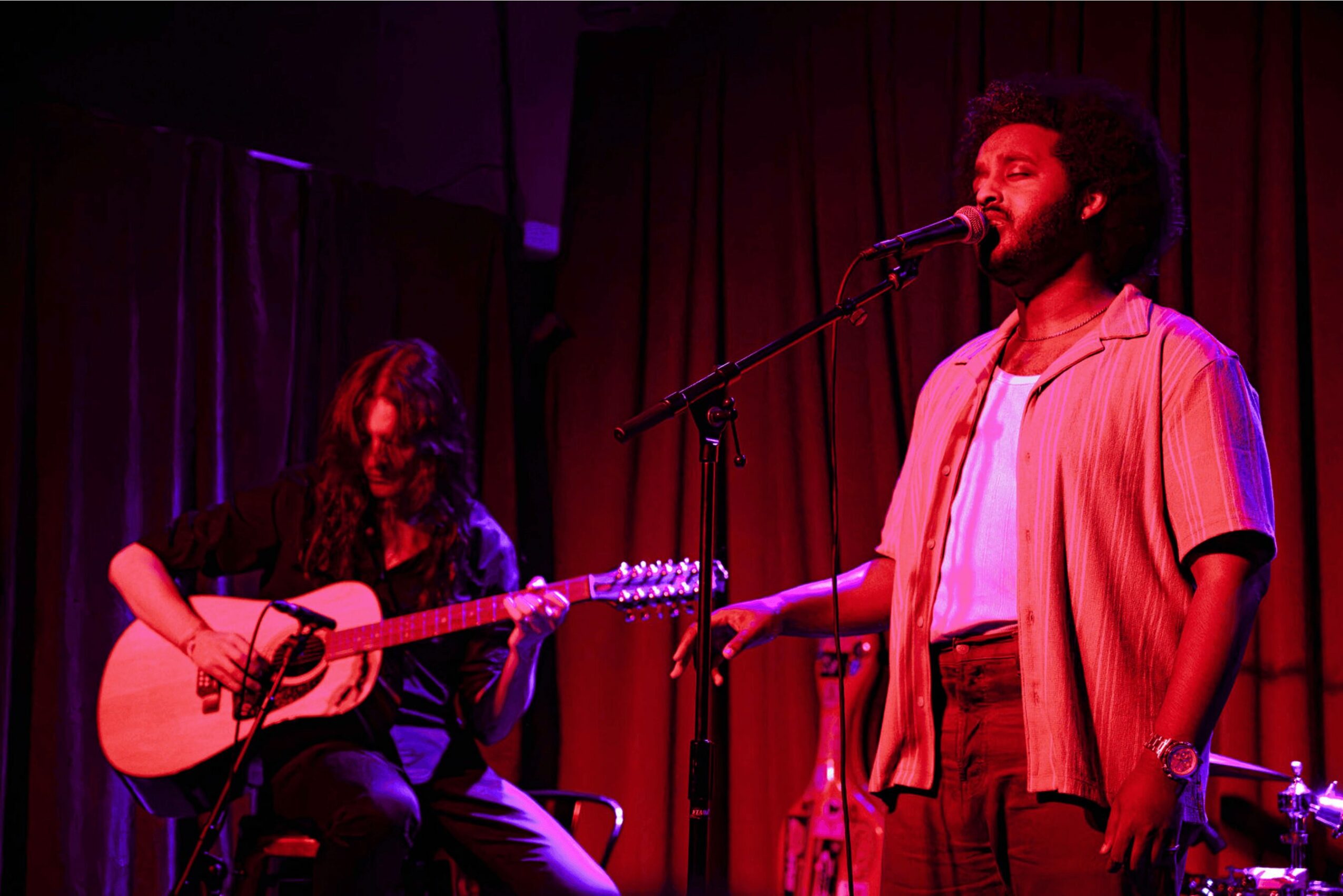 Two people perform on stage, one singing into a microphone and the other playing guitar, with red curtains in the background.