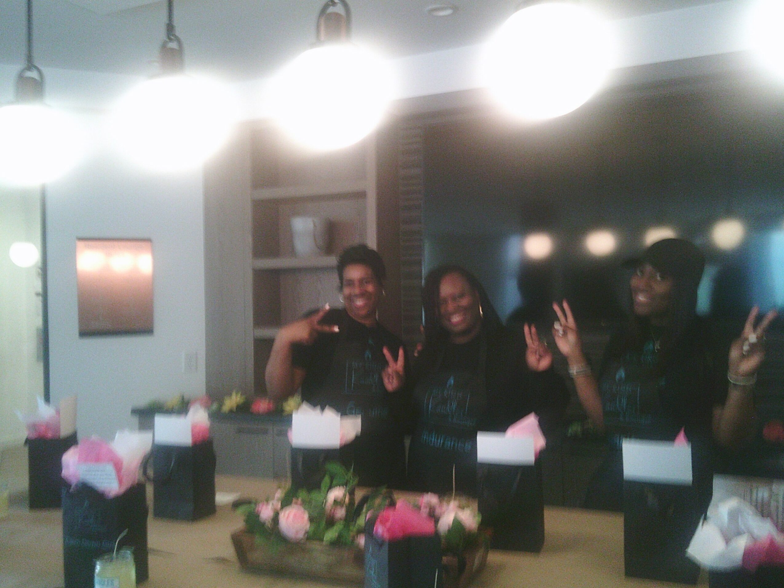 Three women smiling and waving at a table with pink and white flowers, boxes, and a background with lights and shelves.