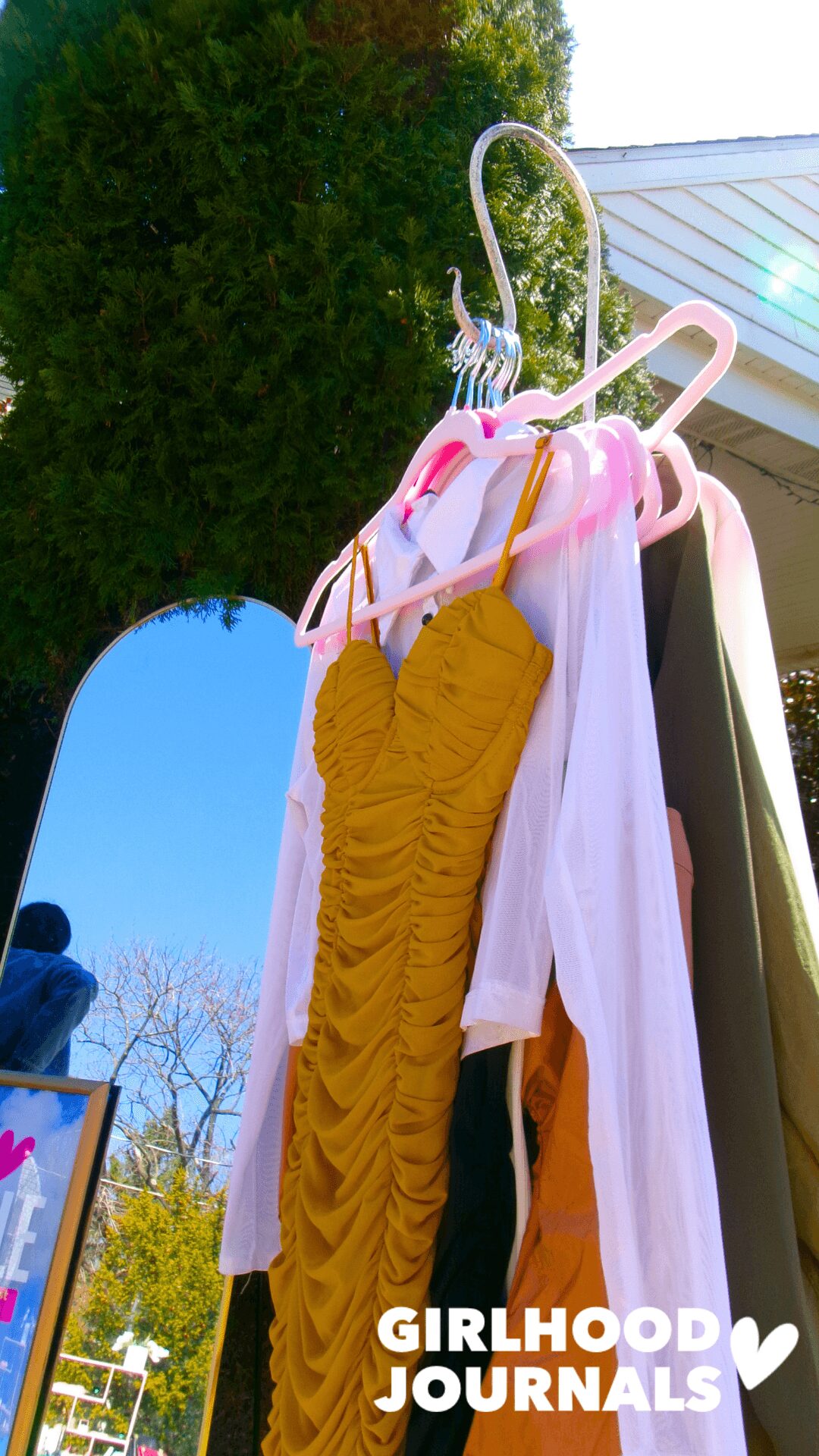 Yellow ruched dress hanging on a pink hanger outdoors with trees and a house in background.
