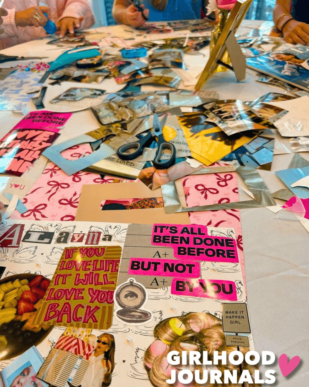 Table covered with photos, scissors, and decorative items, with people around, some holding photos, in a lively gathering.