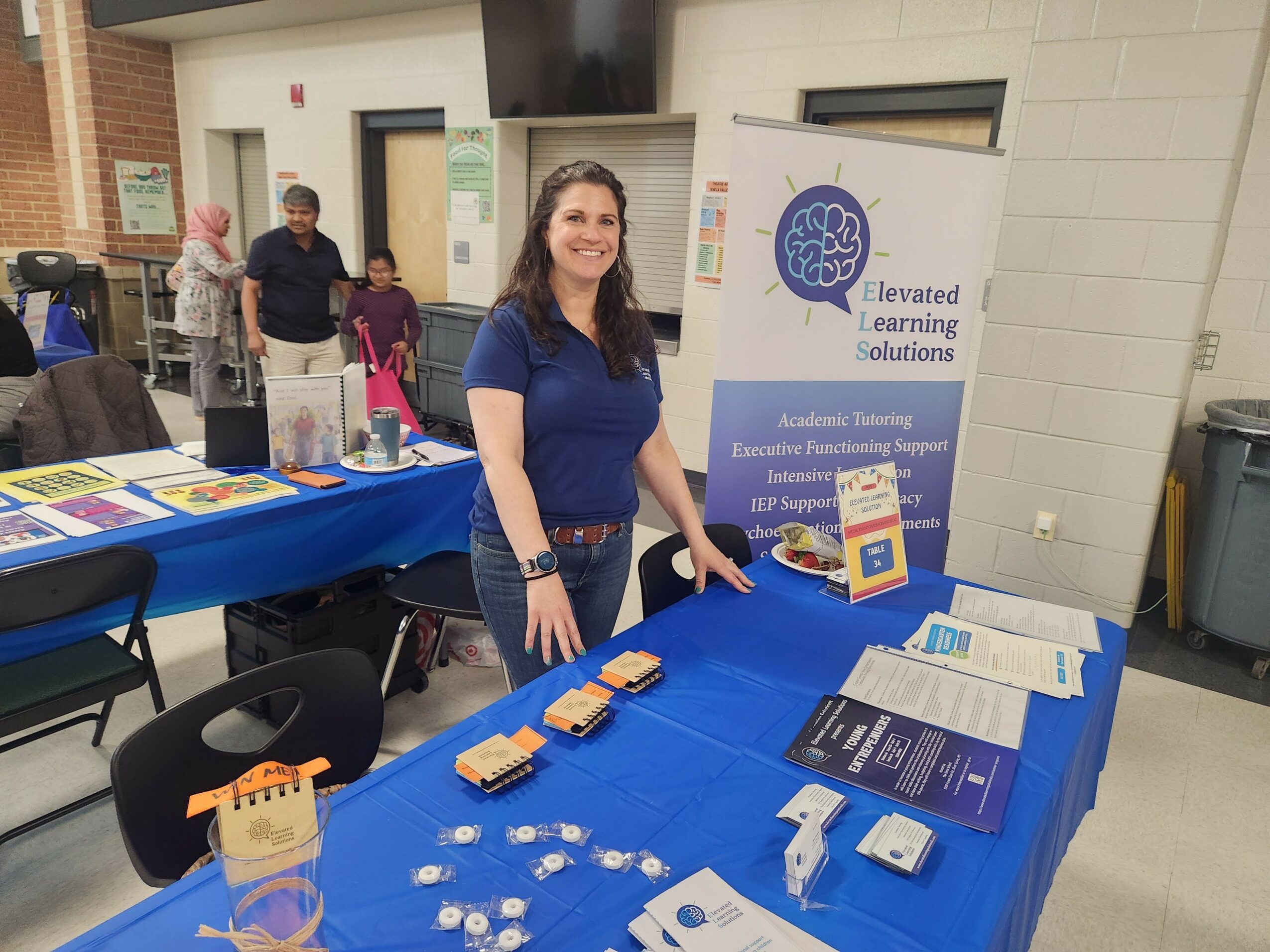 Woman standing behind a table with informational materials at an indoor event, smiling, with a banner in the background.