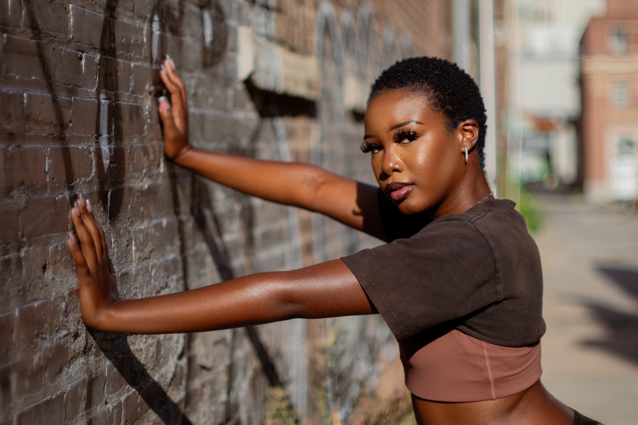 Woman with short hair and dark clothing leaning against a brick wall outdoors, sunlight casting shadows, urban background.
