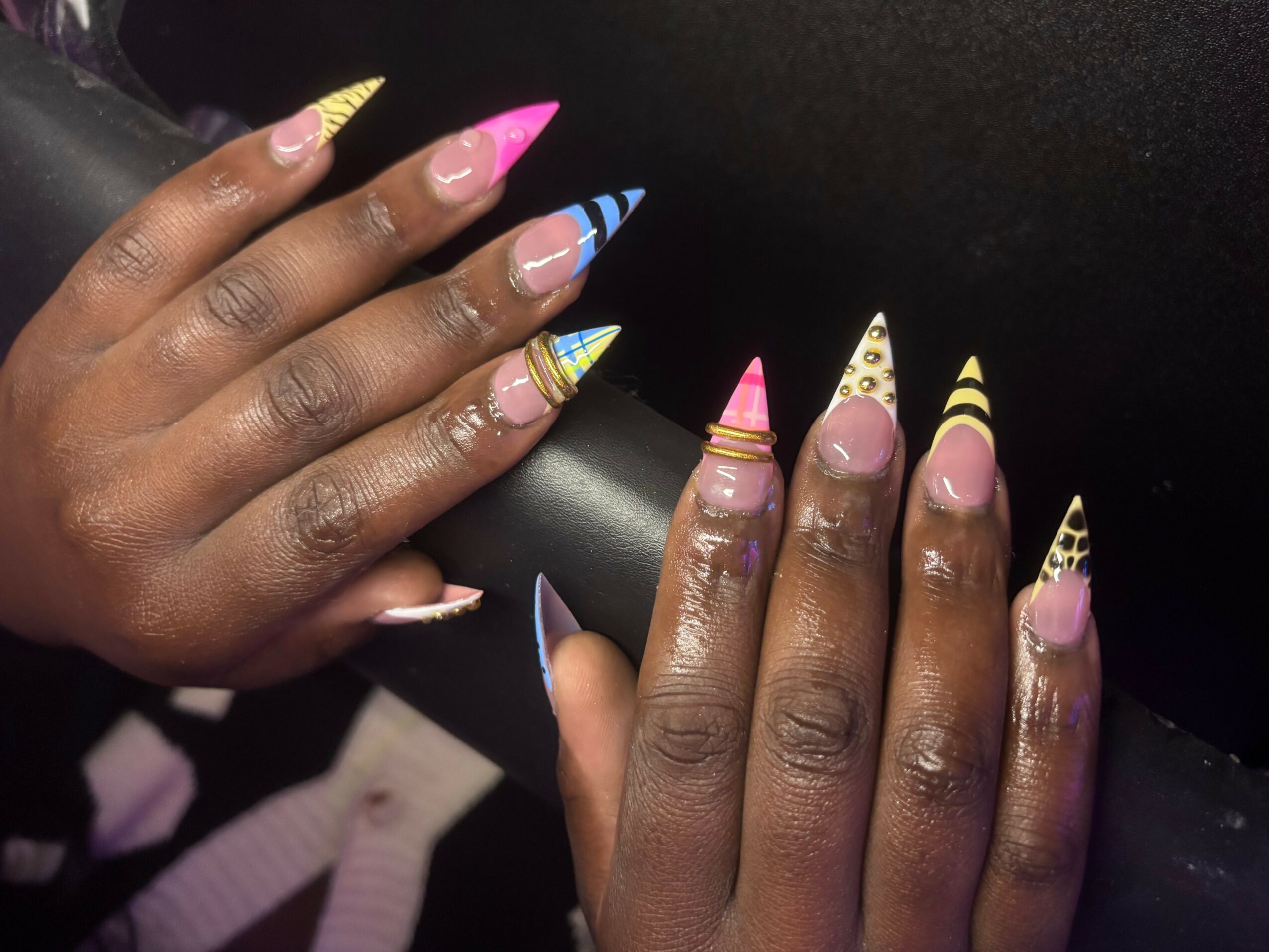 Two hands with colorful, patterned, long, pointed nails resting on a black surface.