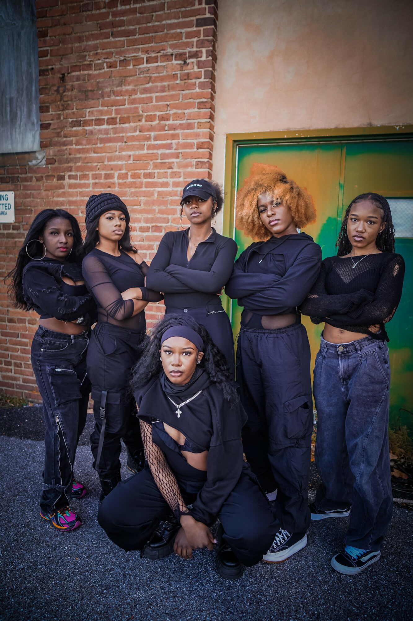 Group of six women standing and sitting against a brick wall and green door, posing confidently outdoors.